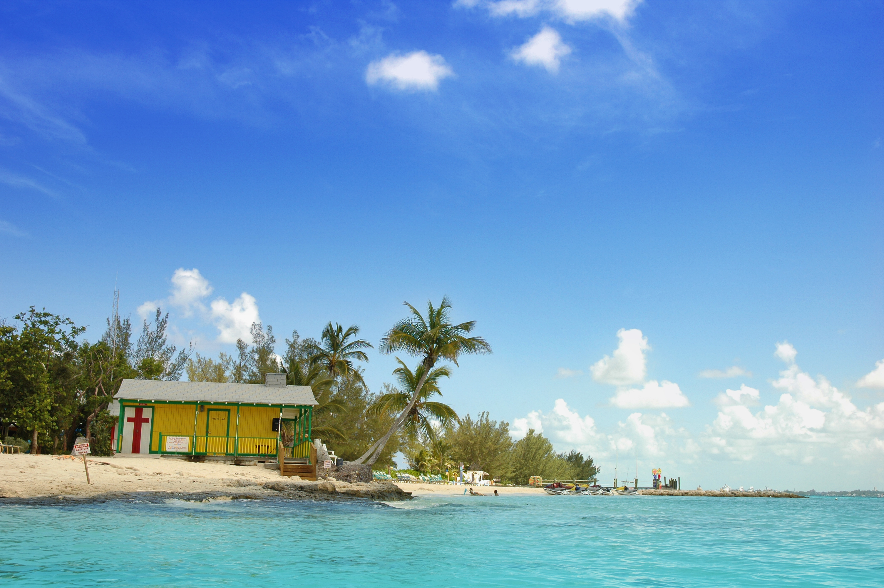 Casa amarilla en playa tropical con palmeras y mar turquesa bajo cielo azul.
