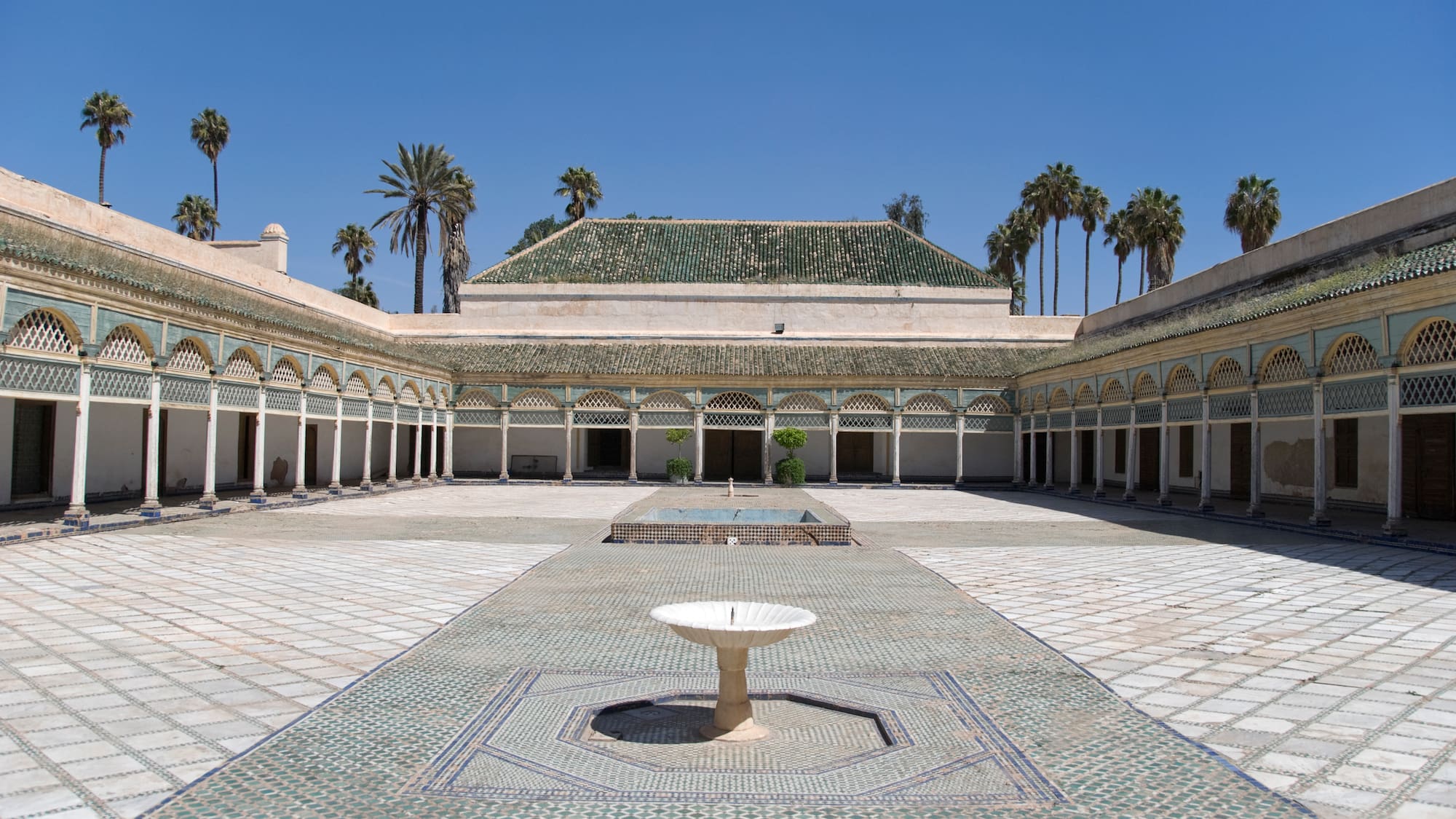 a courtyard with a fountain and palm trees with Bahia Palace in the background