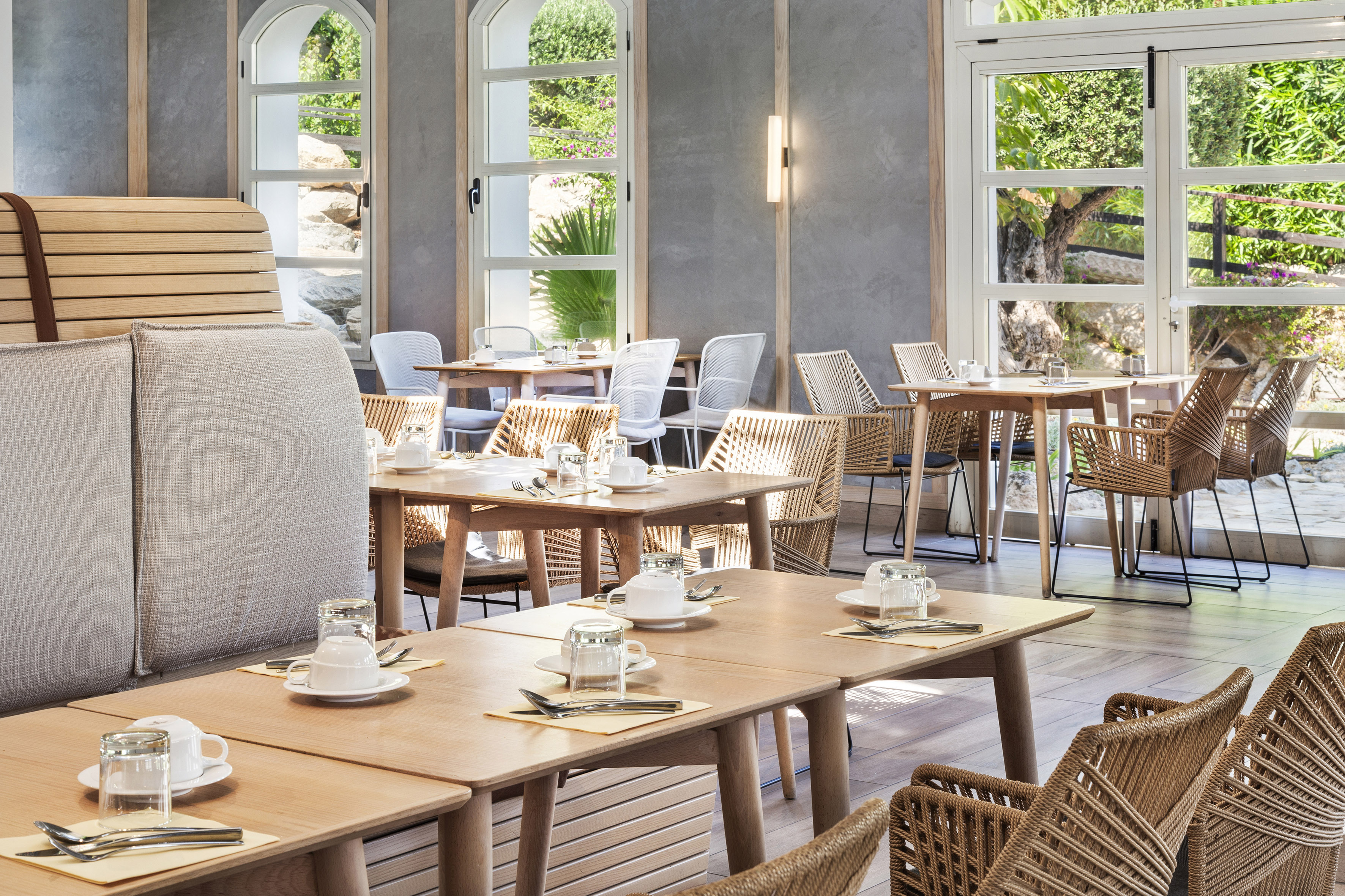 Modern restaurant dining room with wooden tables, woven chairs, and garden views.