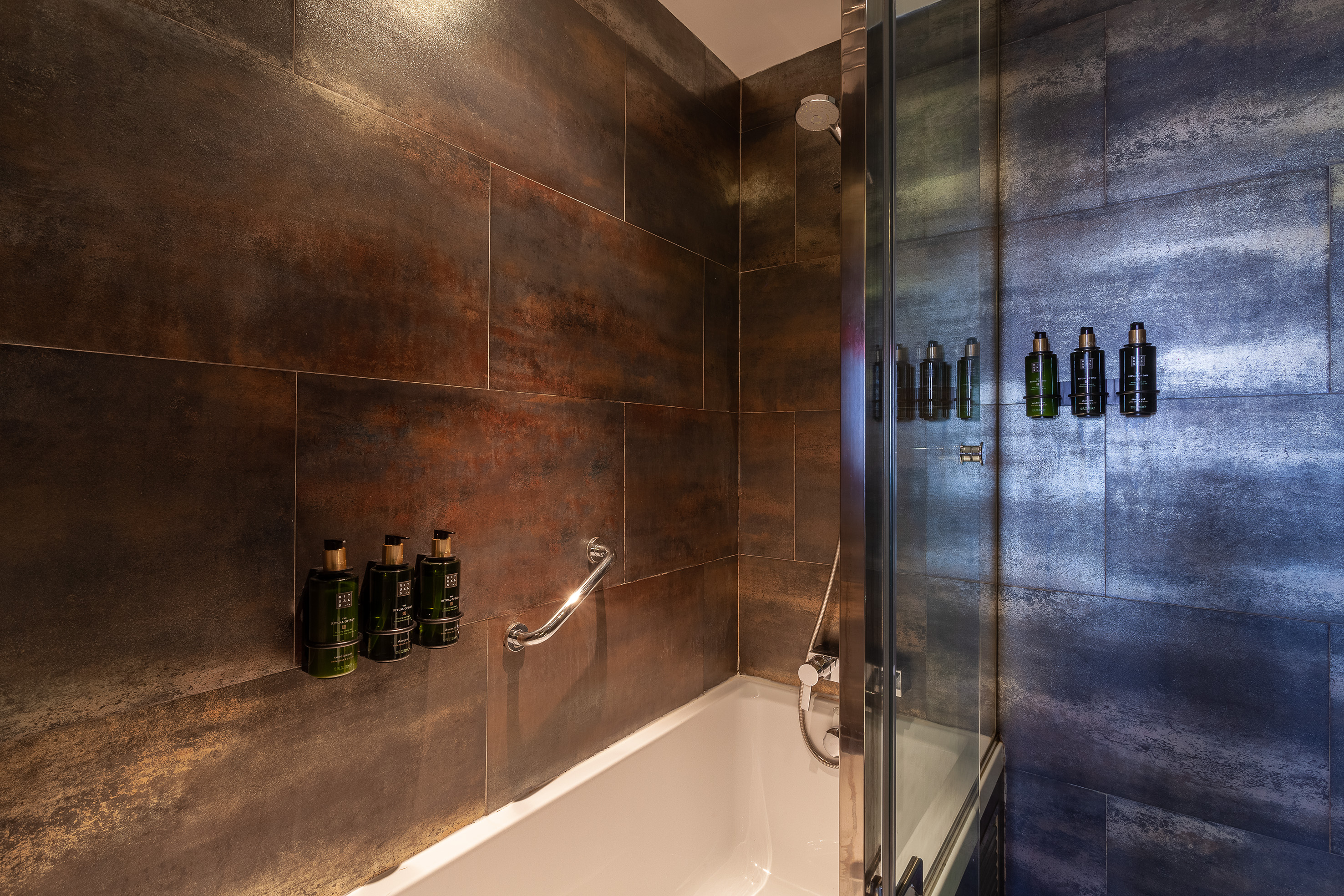 Modern hotel bathroom with dark tiles, bathtub, shower, and toiletries.