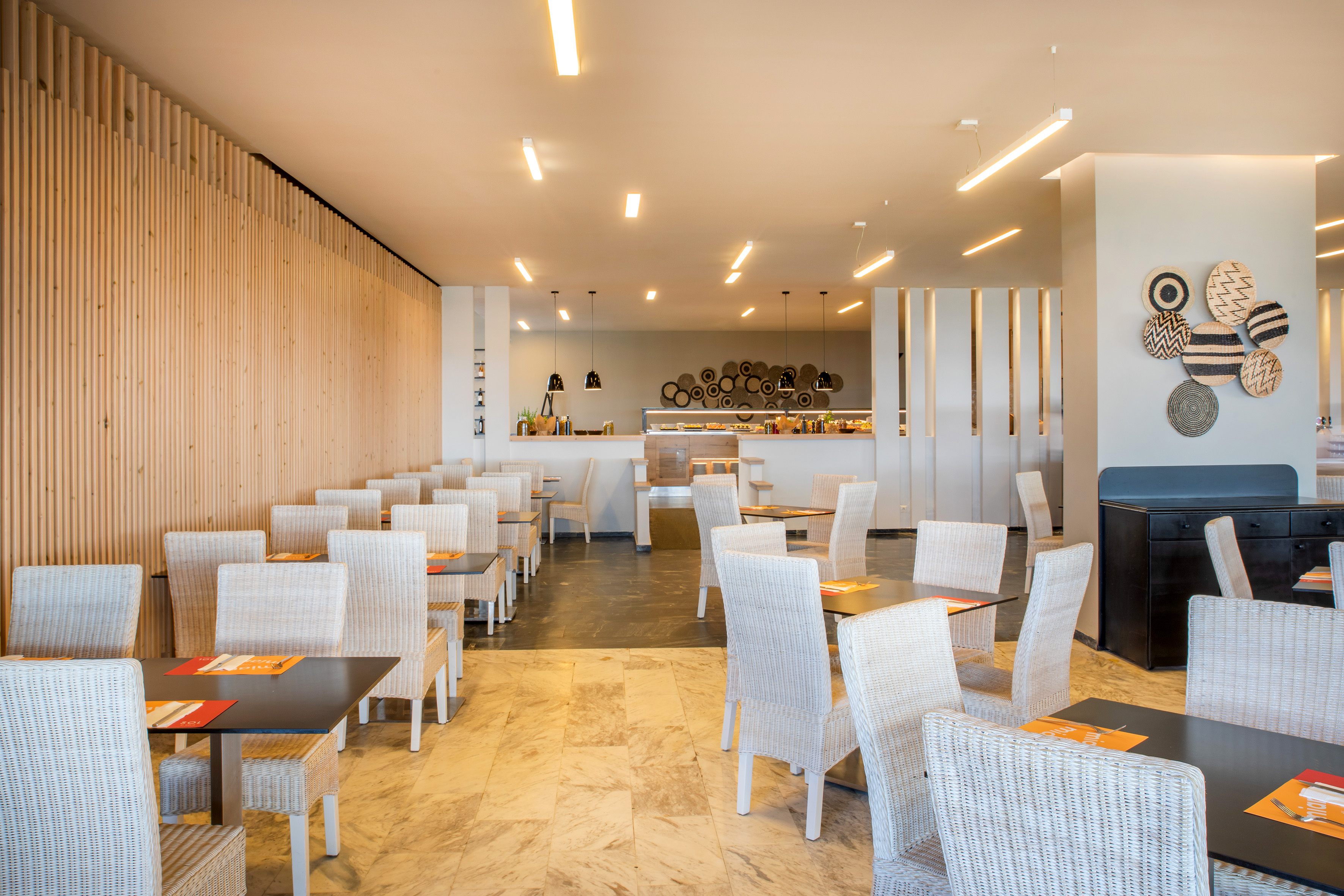 Modern hotel dining room with buffet, light wood wall, and white wicker chairs.