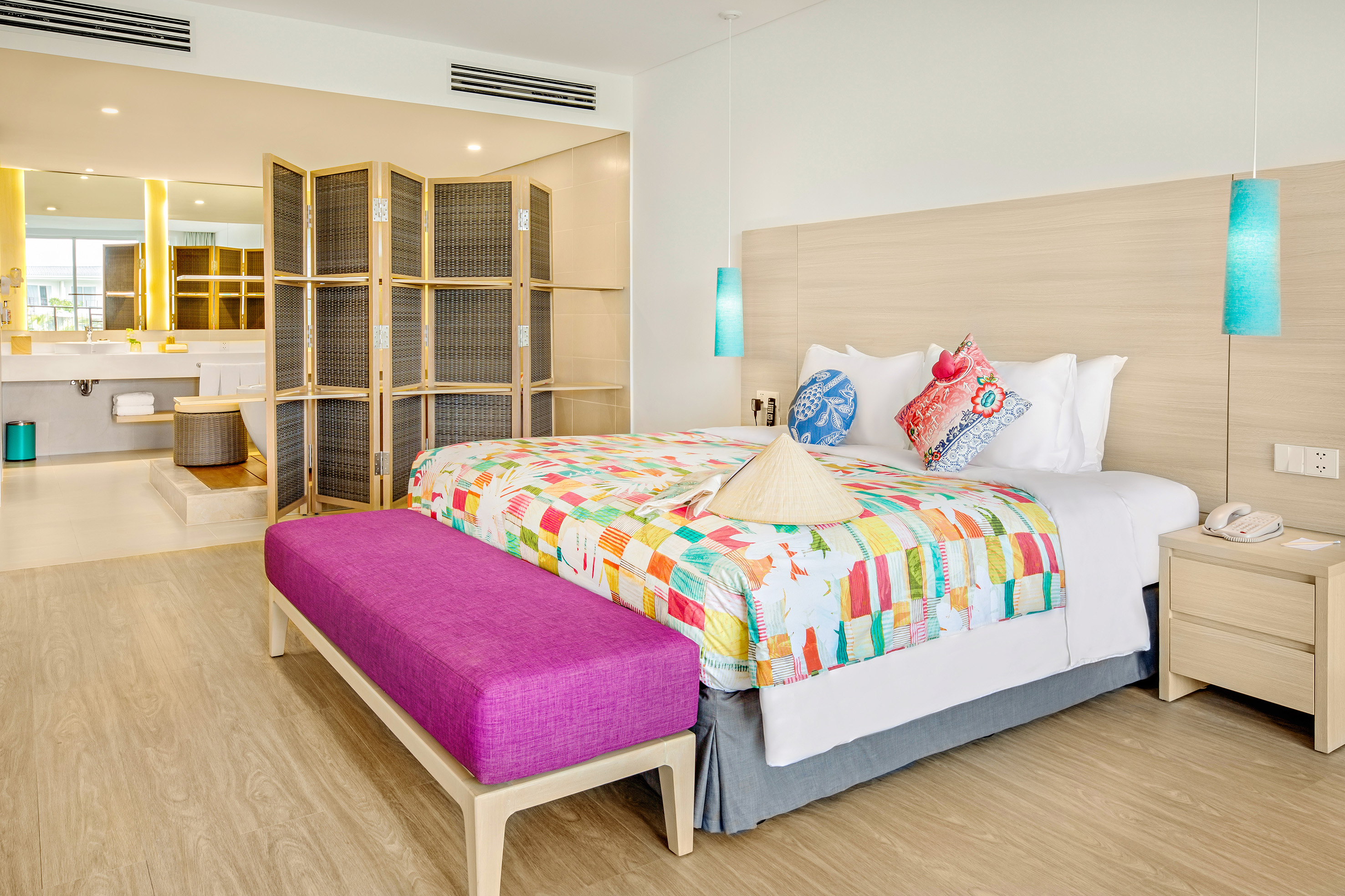 Bright hotel bedroom with colorful quilt, purple bench, and open-plan bathroom.