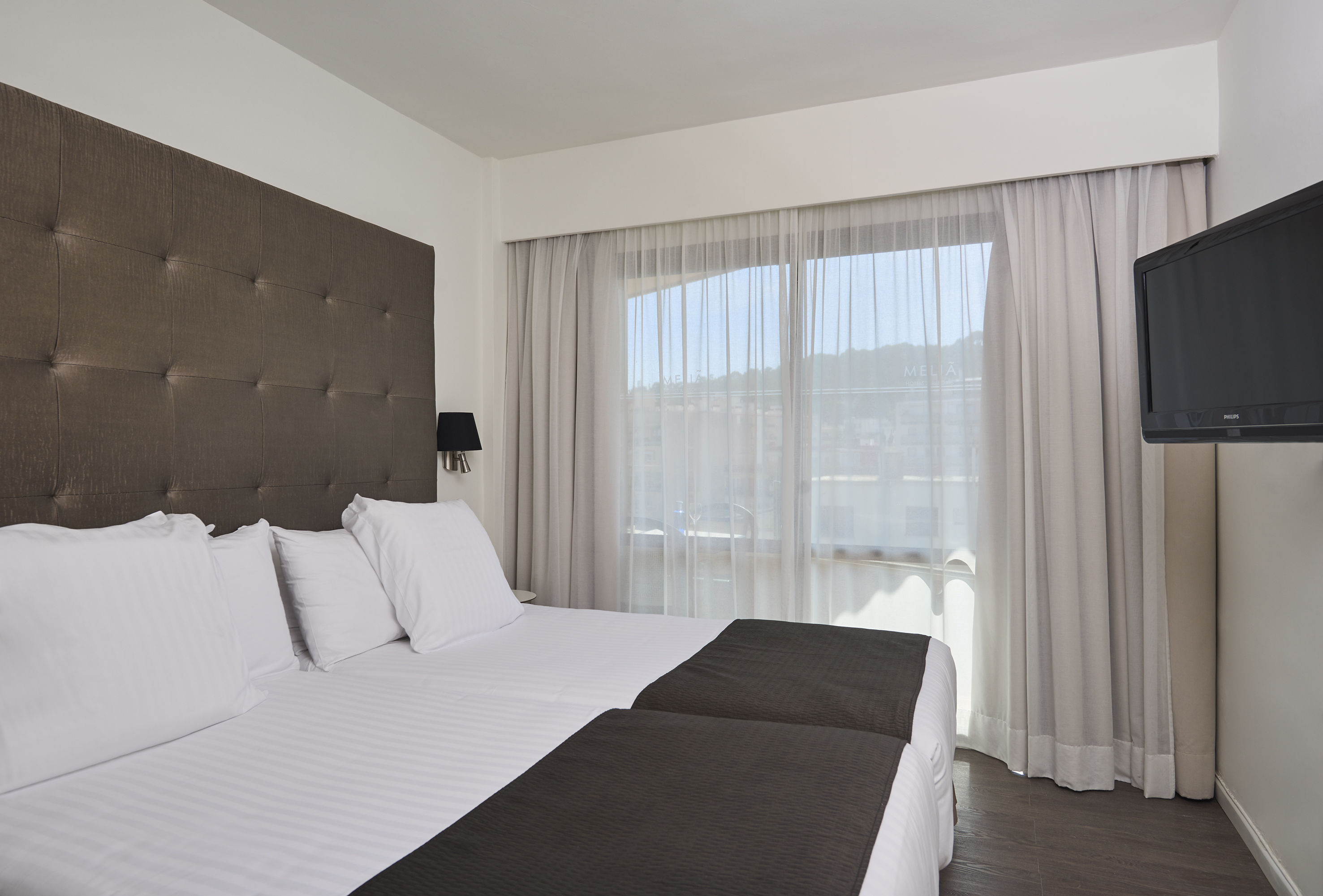 Modern hotel room with twin beds, brown headboard, curtains, and wall-mounted TV.