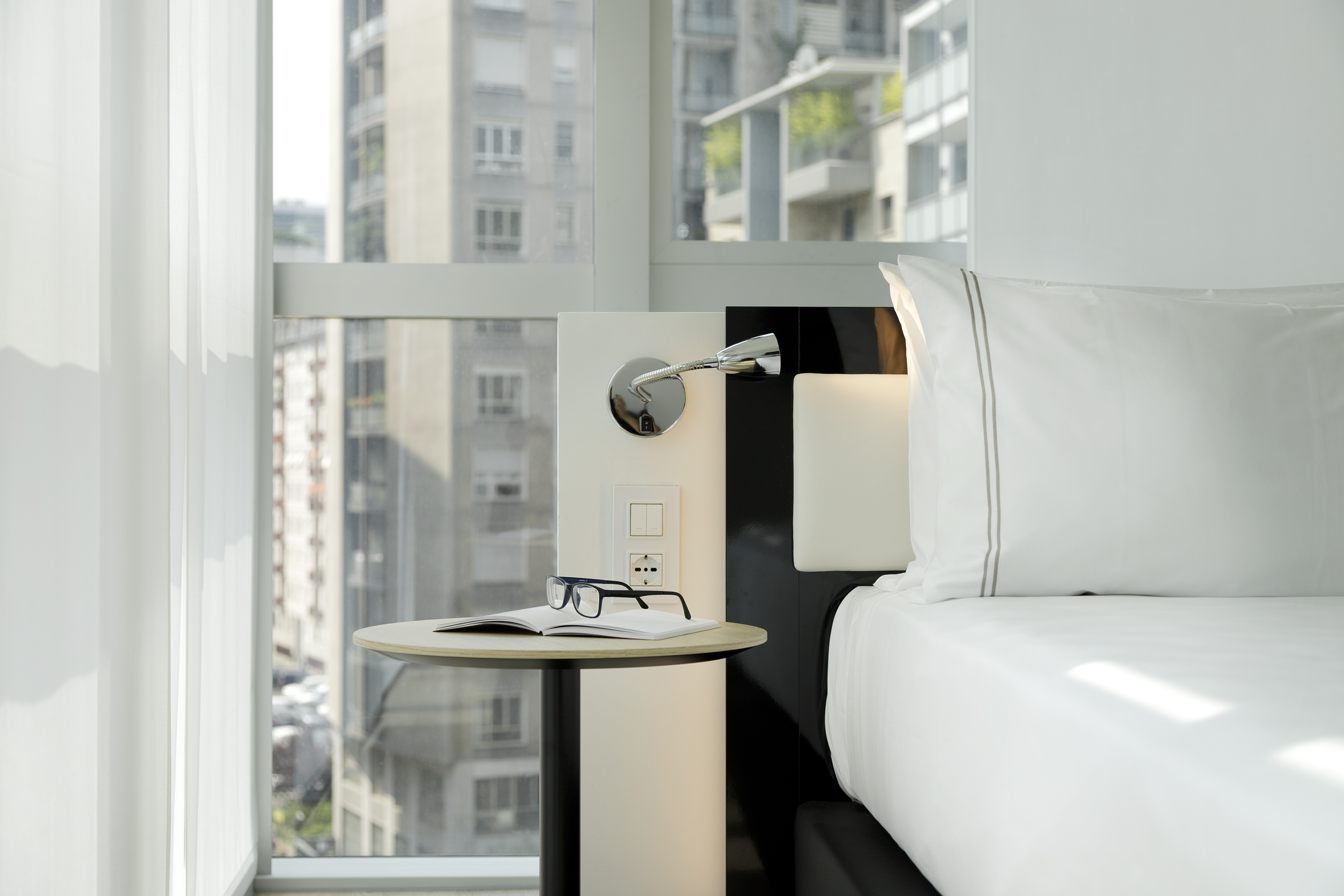 Modern hotel bedroom with white bed, bedside table, book, and city view.
