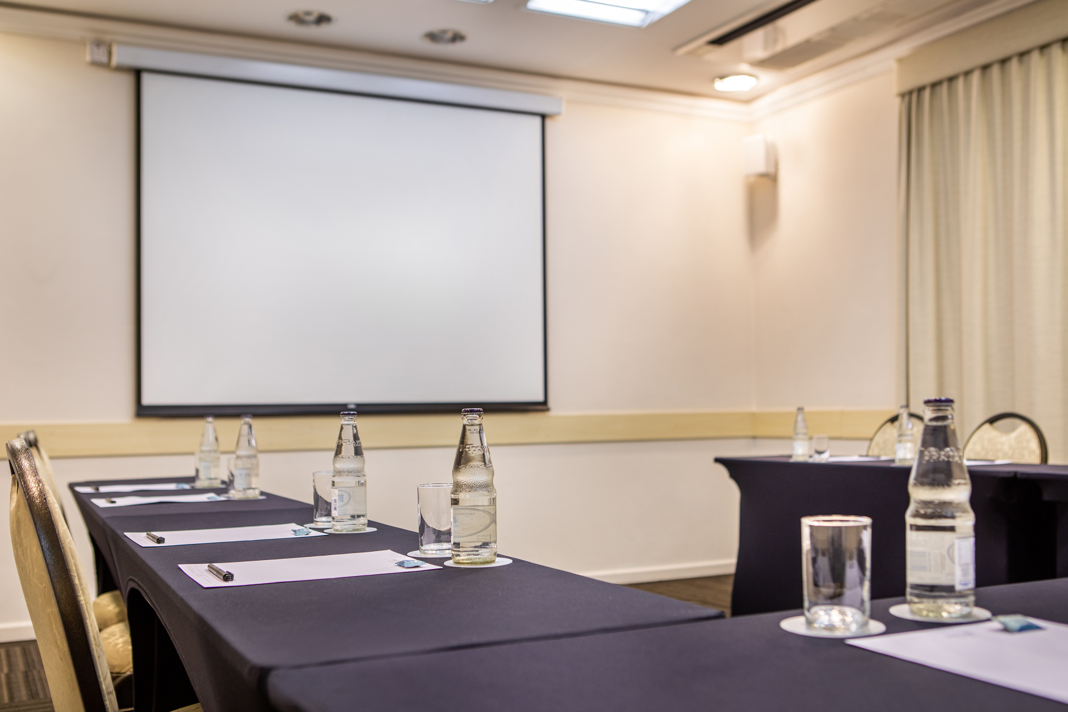 Empty meeting room with projector screen, tables, water bottles, and notepads.