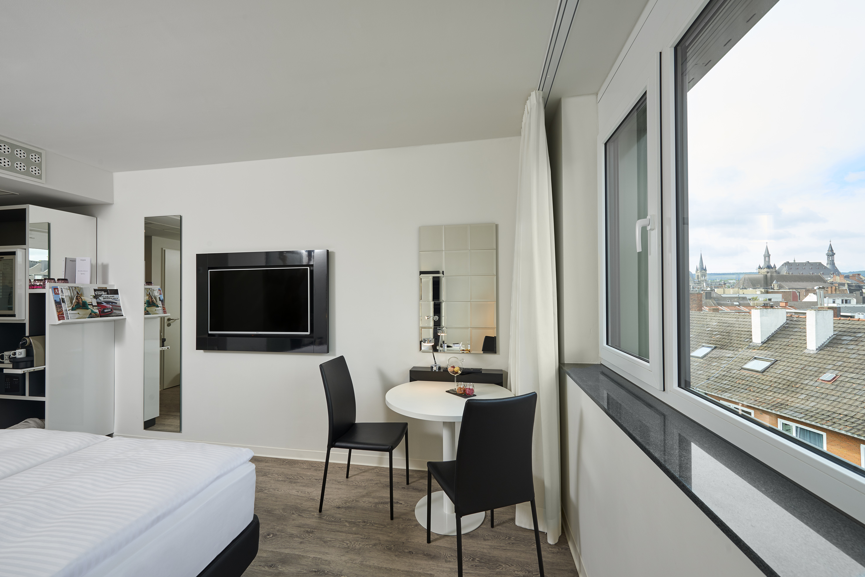 Modern hotel room with city view, comfortable bed, table, and flat-screen TV.