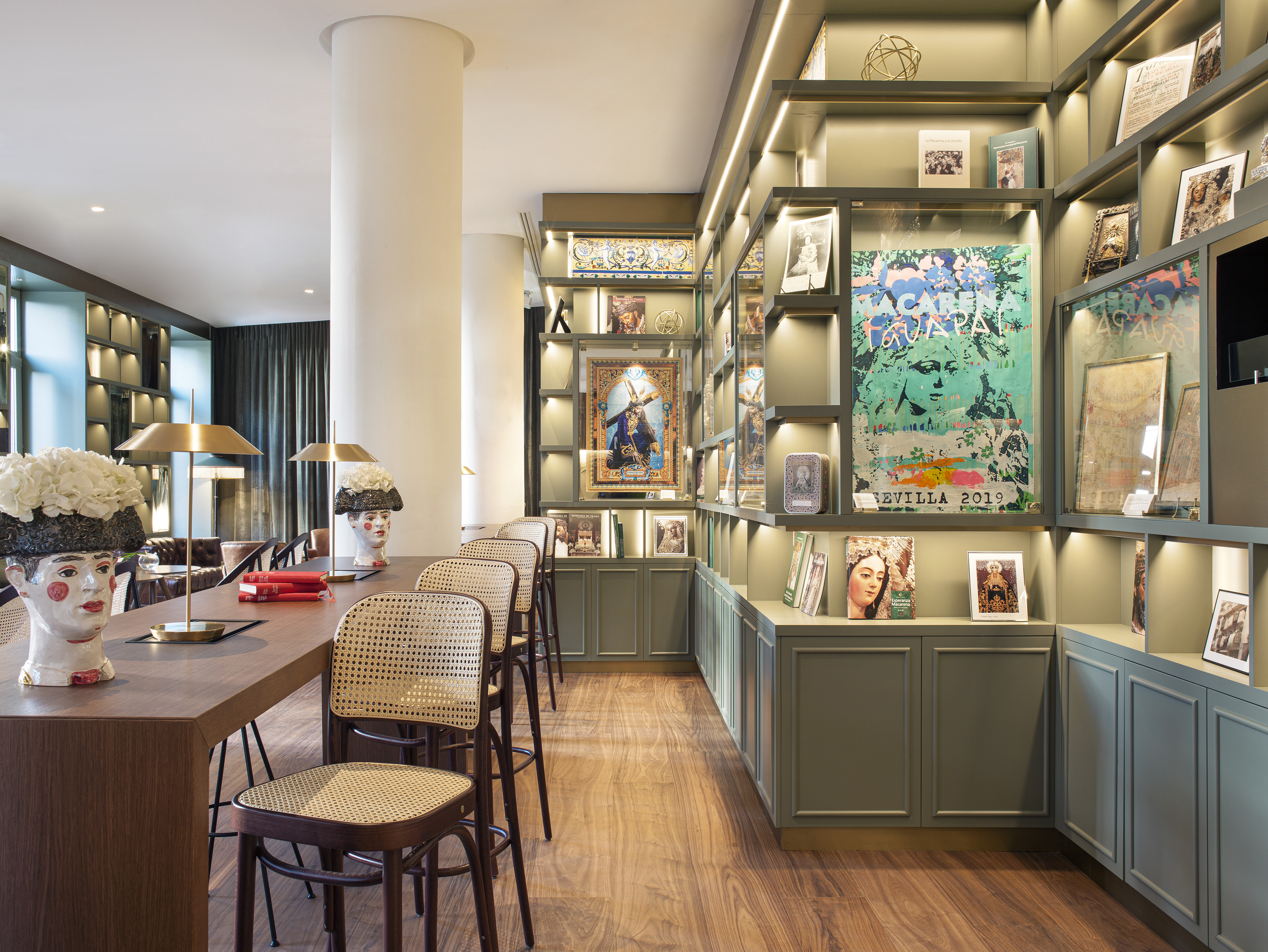 Hotel lounge with long wooden table, cane chairs, and lit green display shelves.