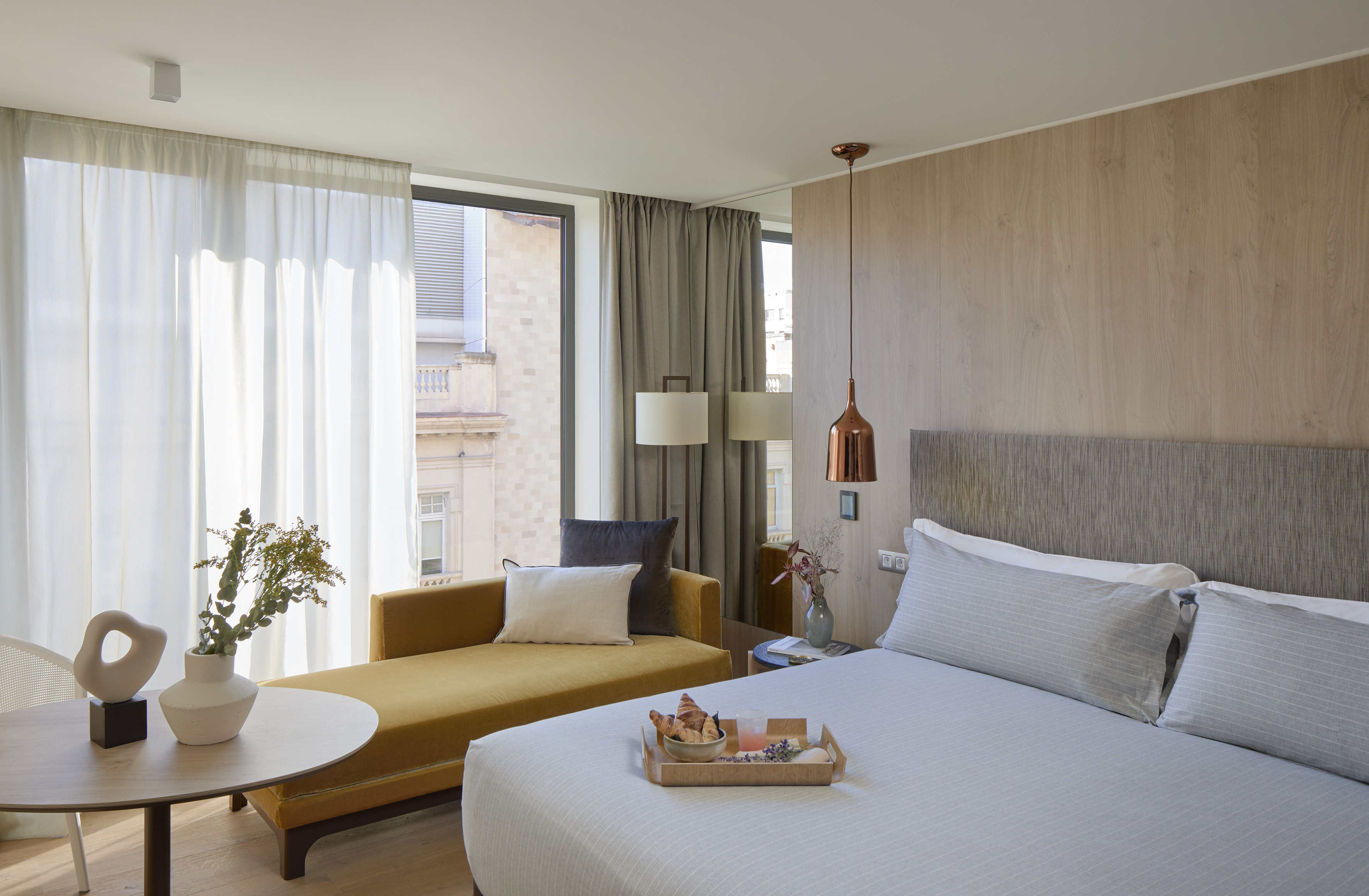 Modern hotel room with breakfast in bed, yellow daybed, and large window.