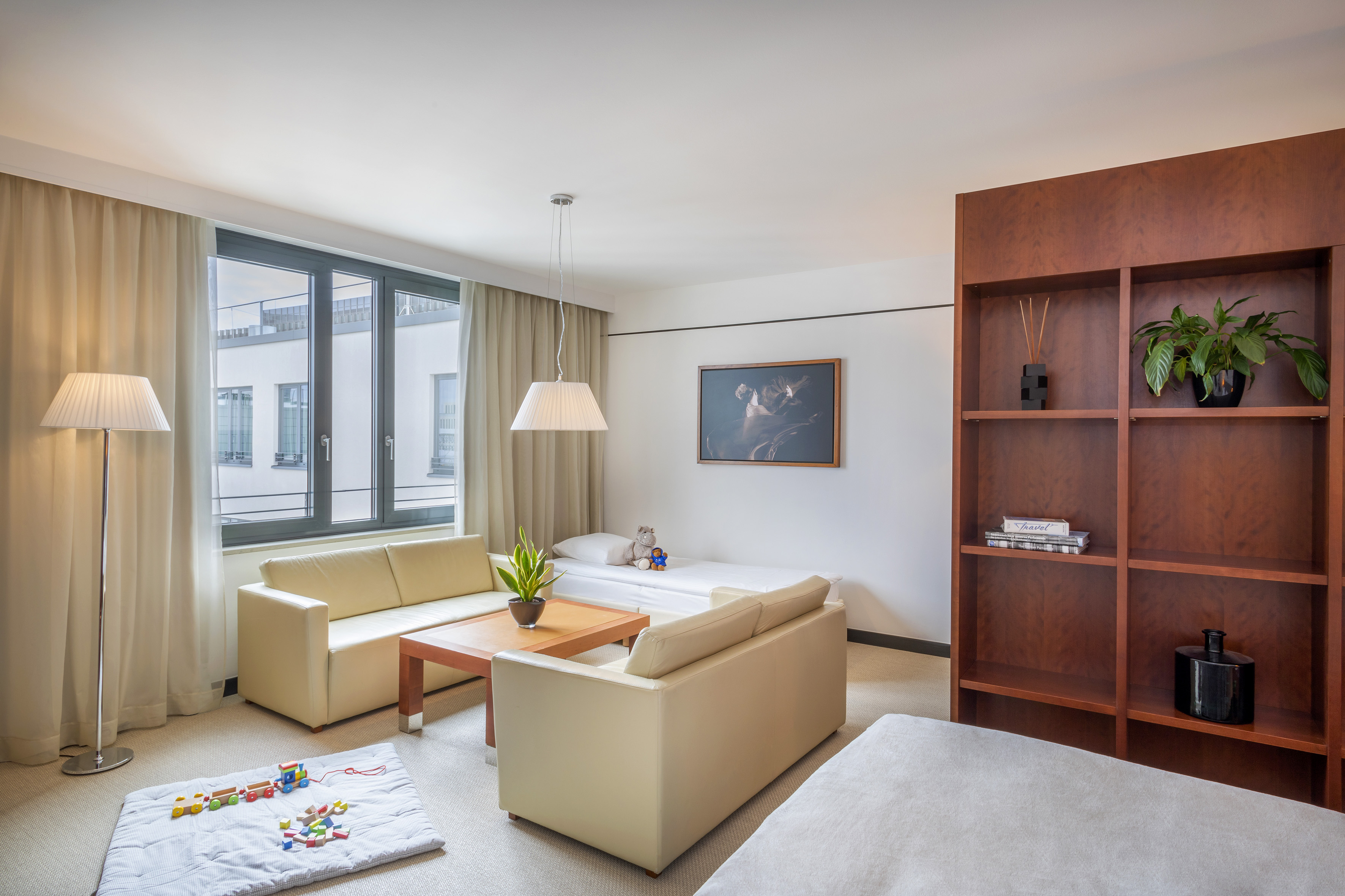 Modern hotel family suite with light sofas, window, wooden shelves, and playmat.