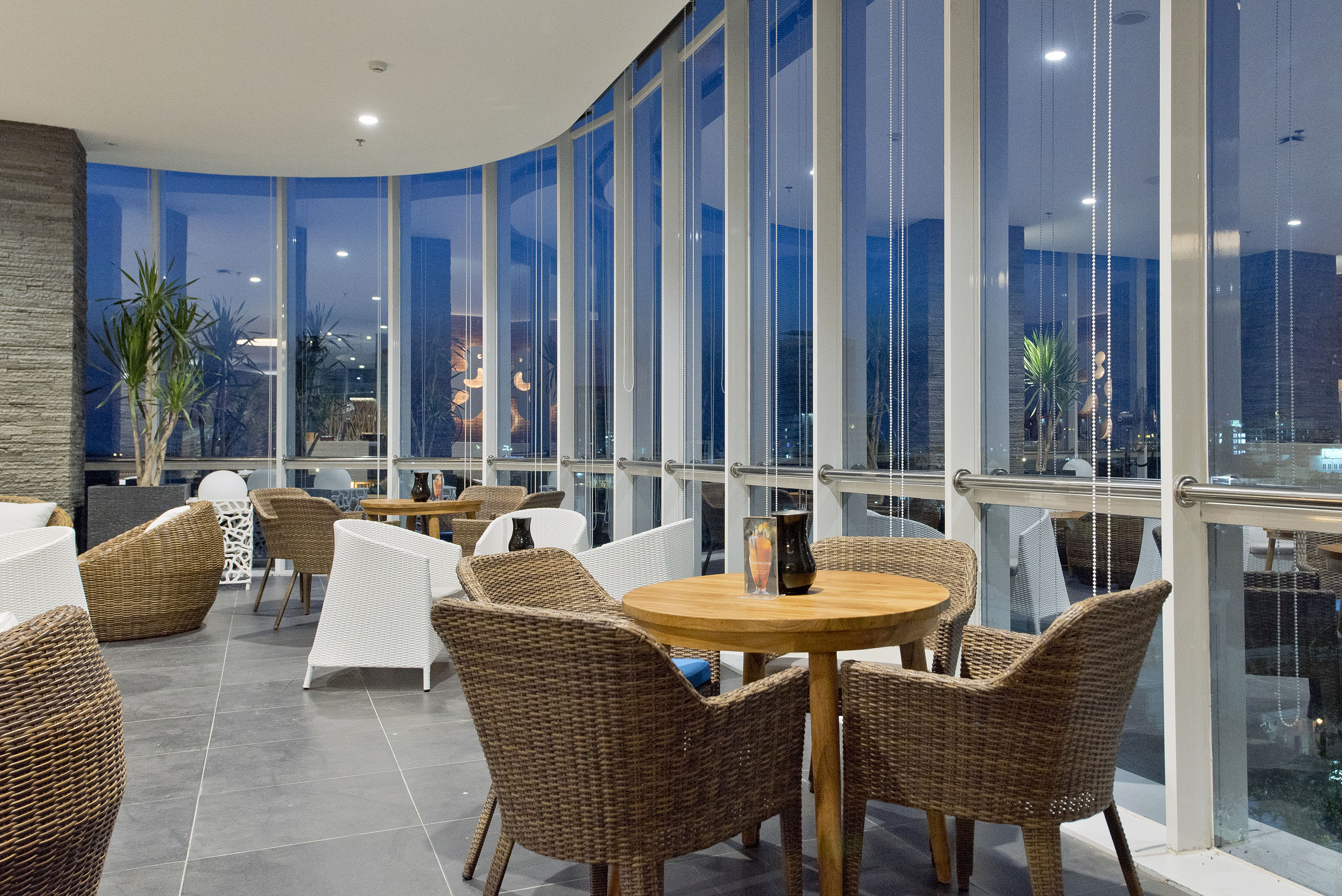 Modern lounge with wicker chairs, wood tables, and curved glass windows at night.