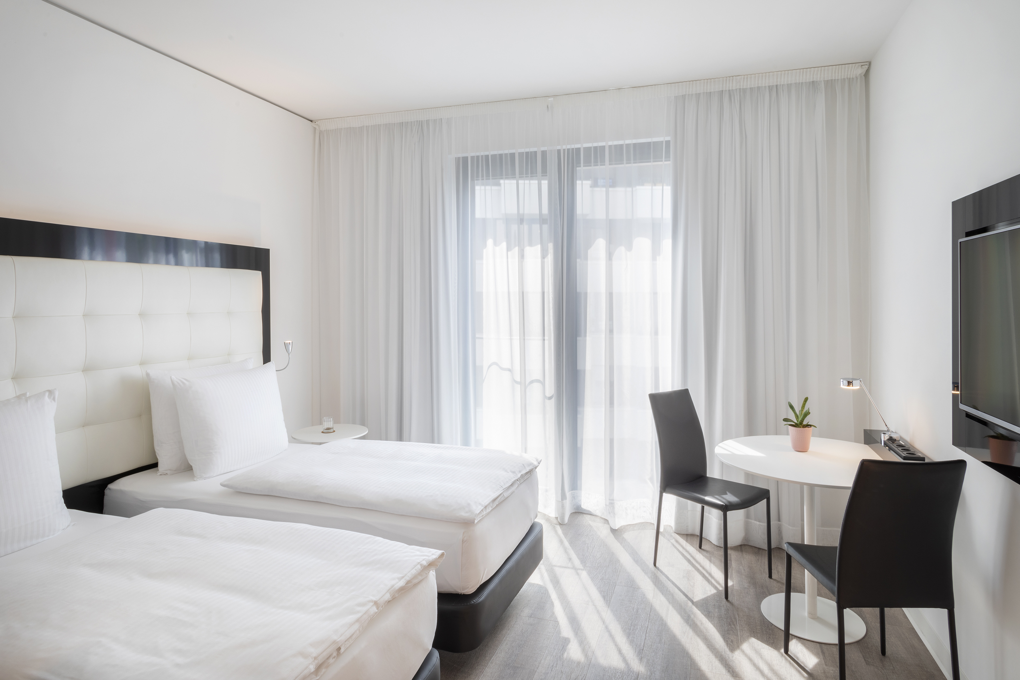 Bright modern hotel room with twin beds, white decor, table, and sunlit window.