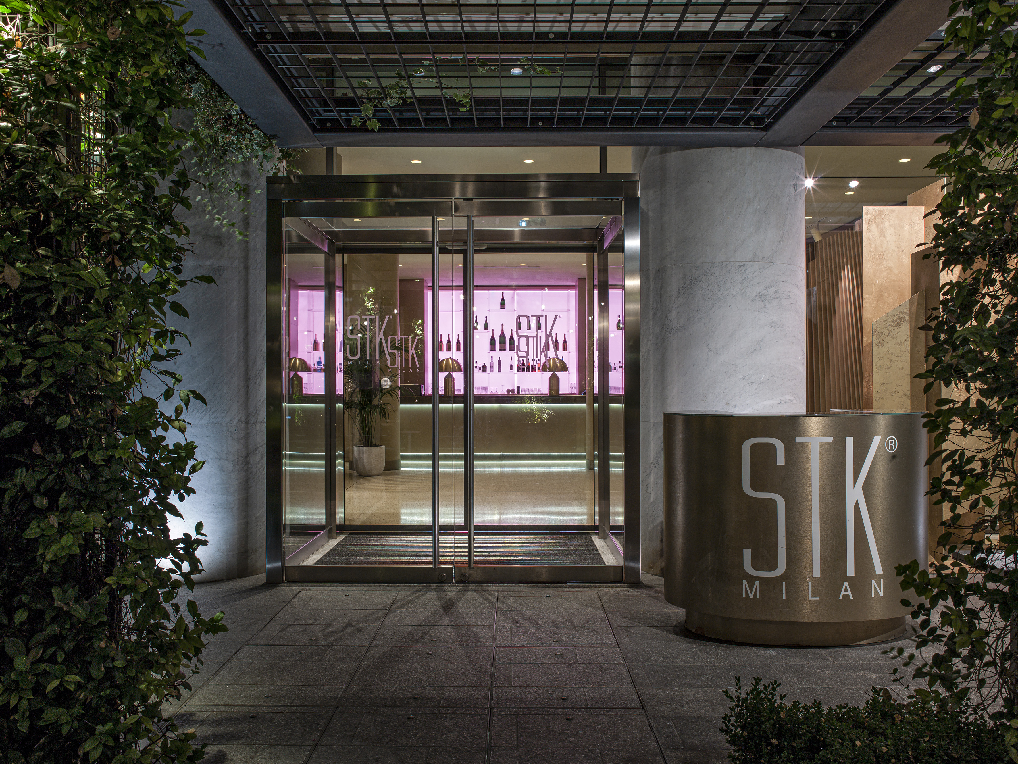 Modern STK Milan restaurant facade at night, glass entrance, pink bar visible inside.