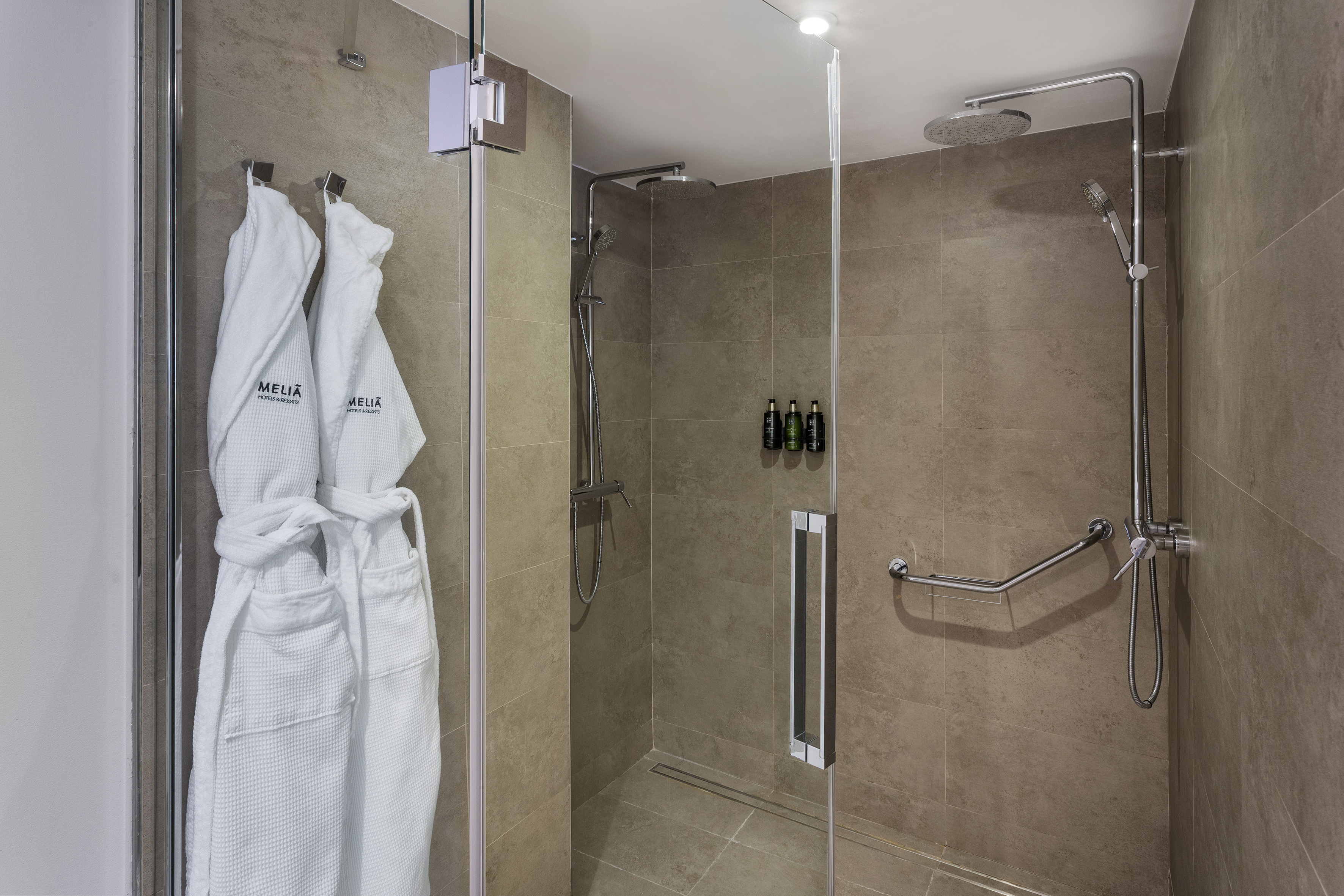 Melia hotel bathroom shower with dual heads, bathrobes, and accessibility grab bar.