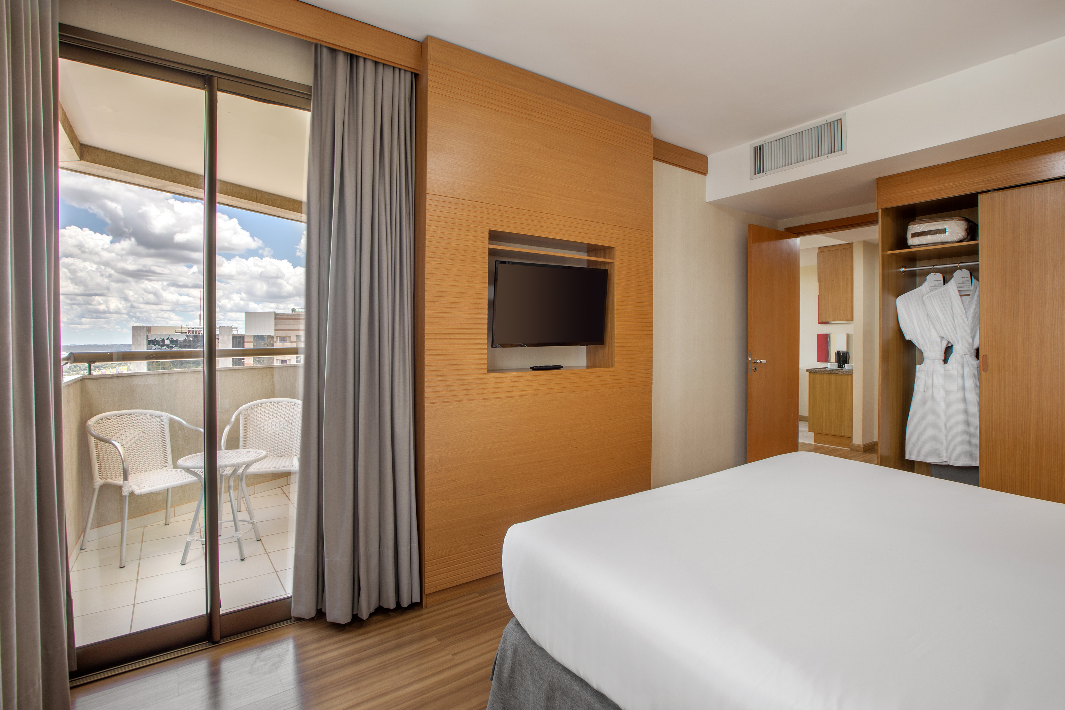 Modern hotel bedroom with balcony, TV, bed, and open closet with bathrobe.