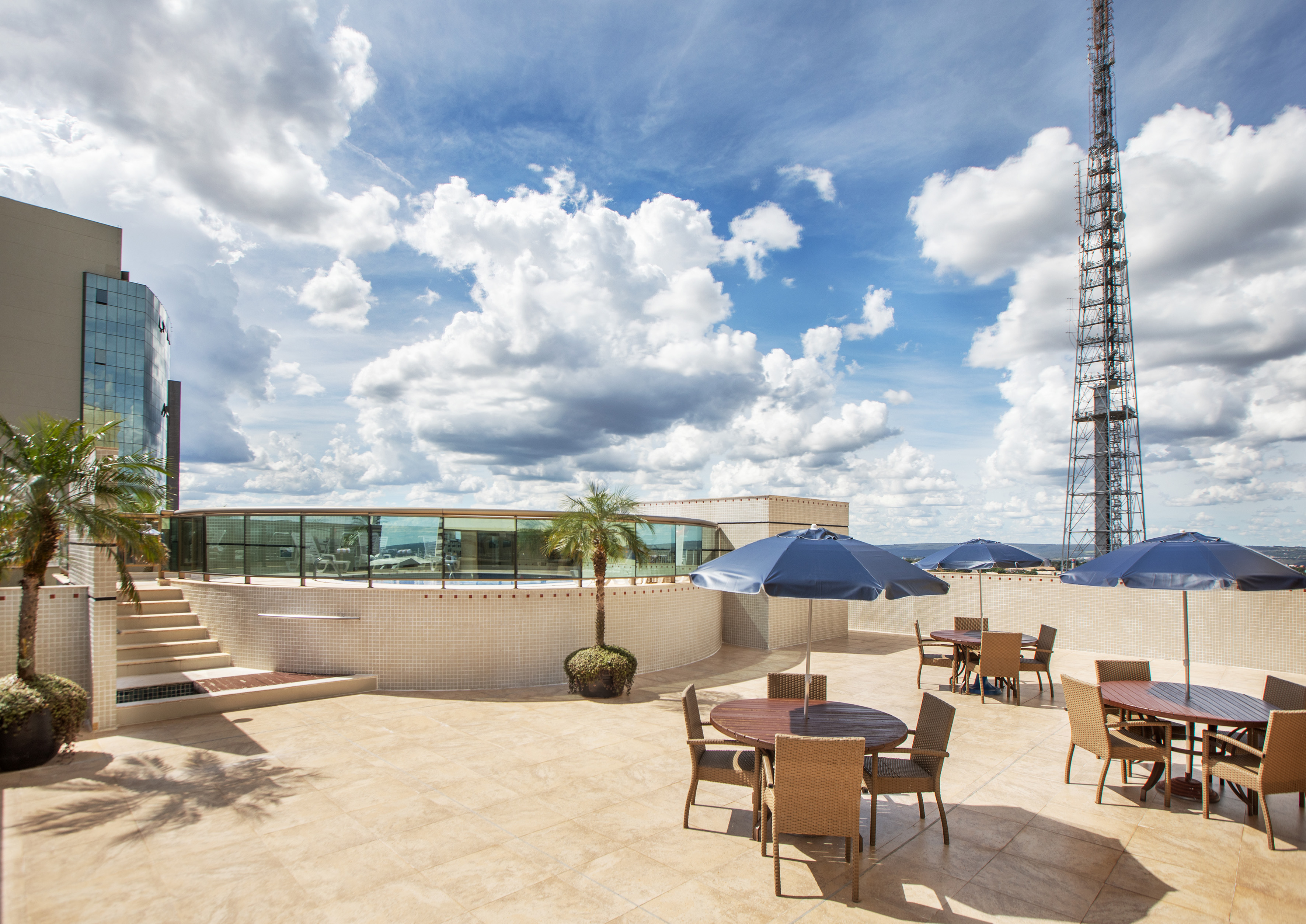 Rooftop terrace with outdoor tables, blue umbrellas, circular pool, and a tall tower.