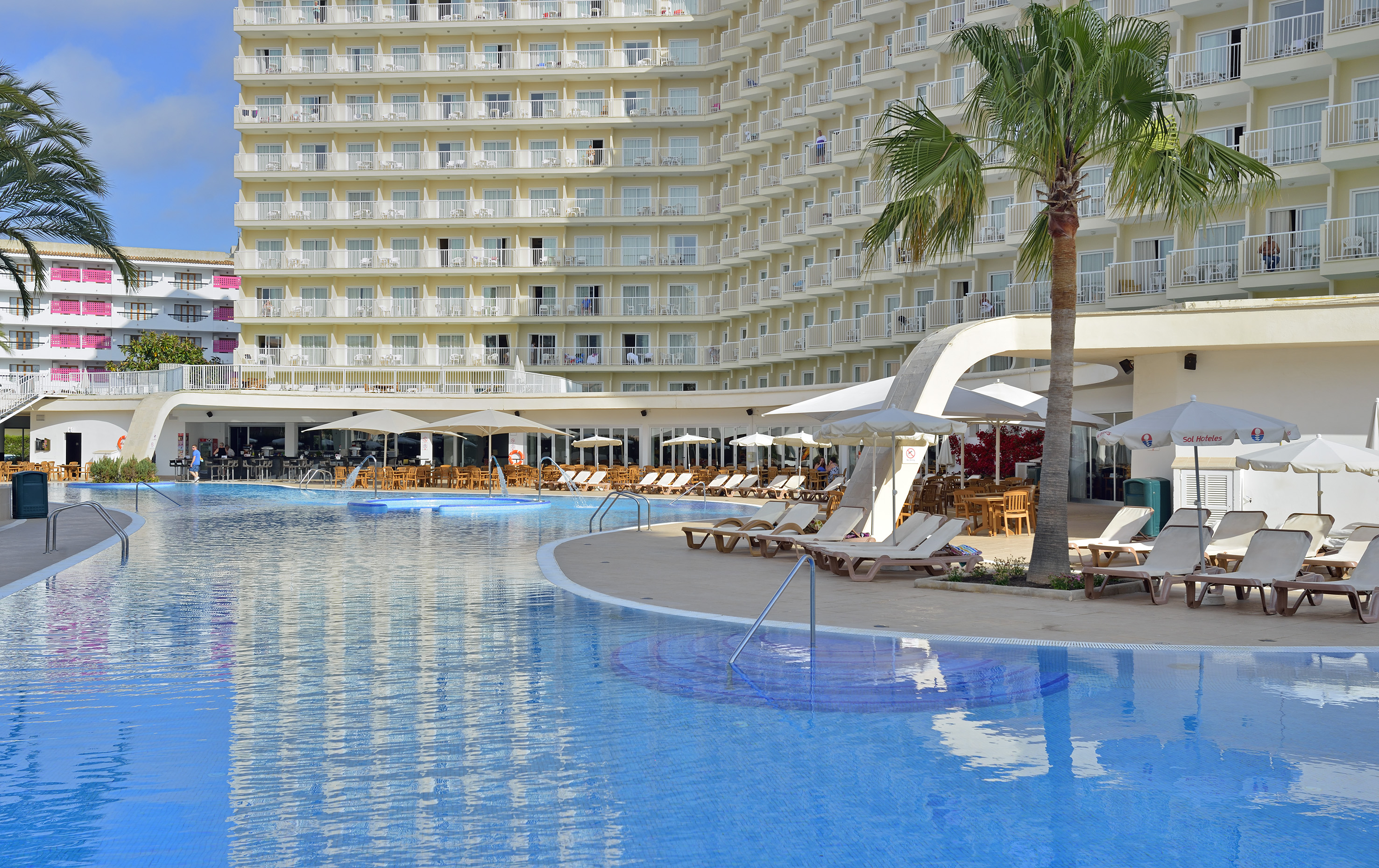 Resort hotel swimming pool, sun loungers, palm trees, and poolside bar.