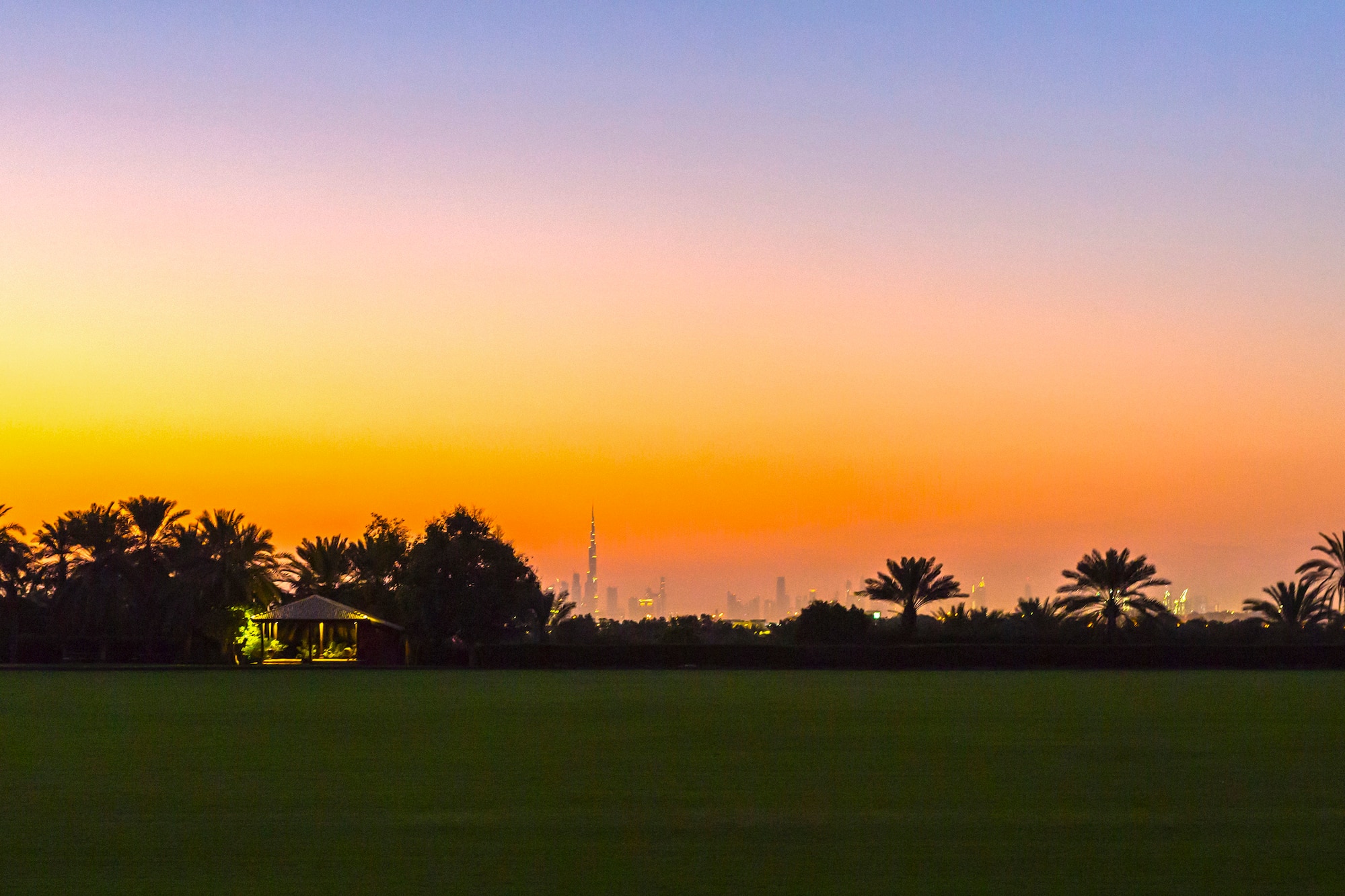 Sunset over Dubai skyline, Burj Khalifa, palm trees, and green field.