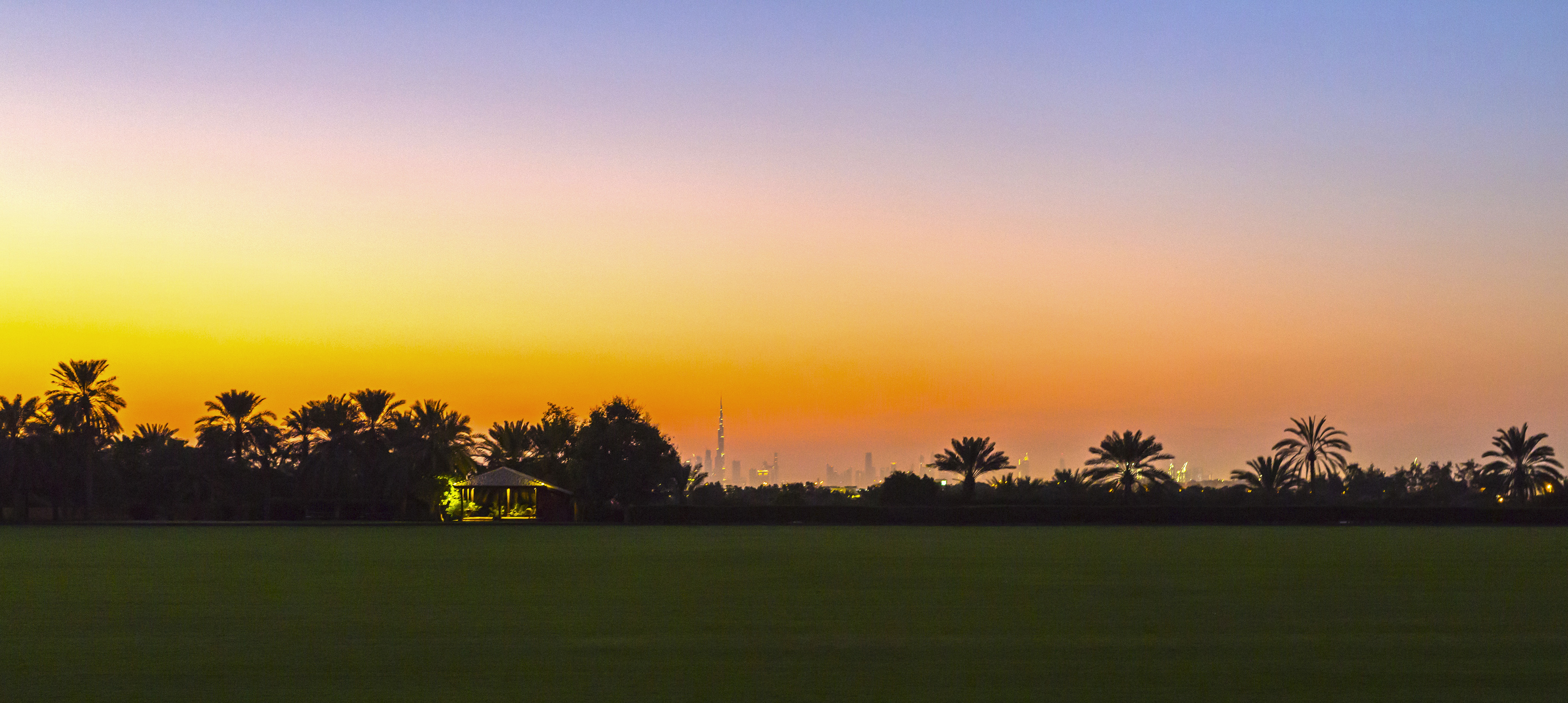 Sunset over Dubai skyline, Burj Khalifa, palm trees, and green field.