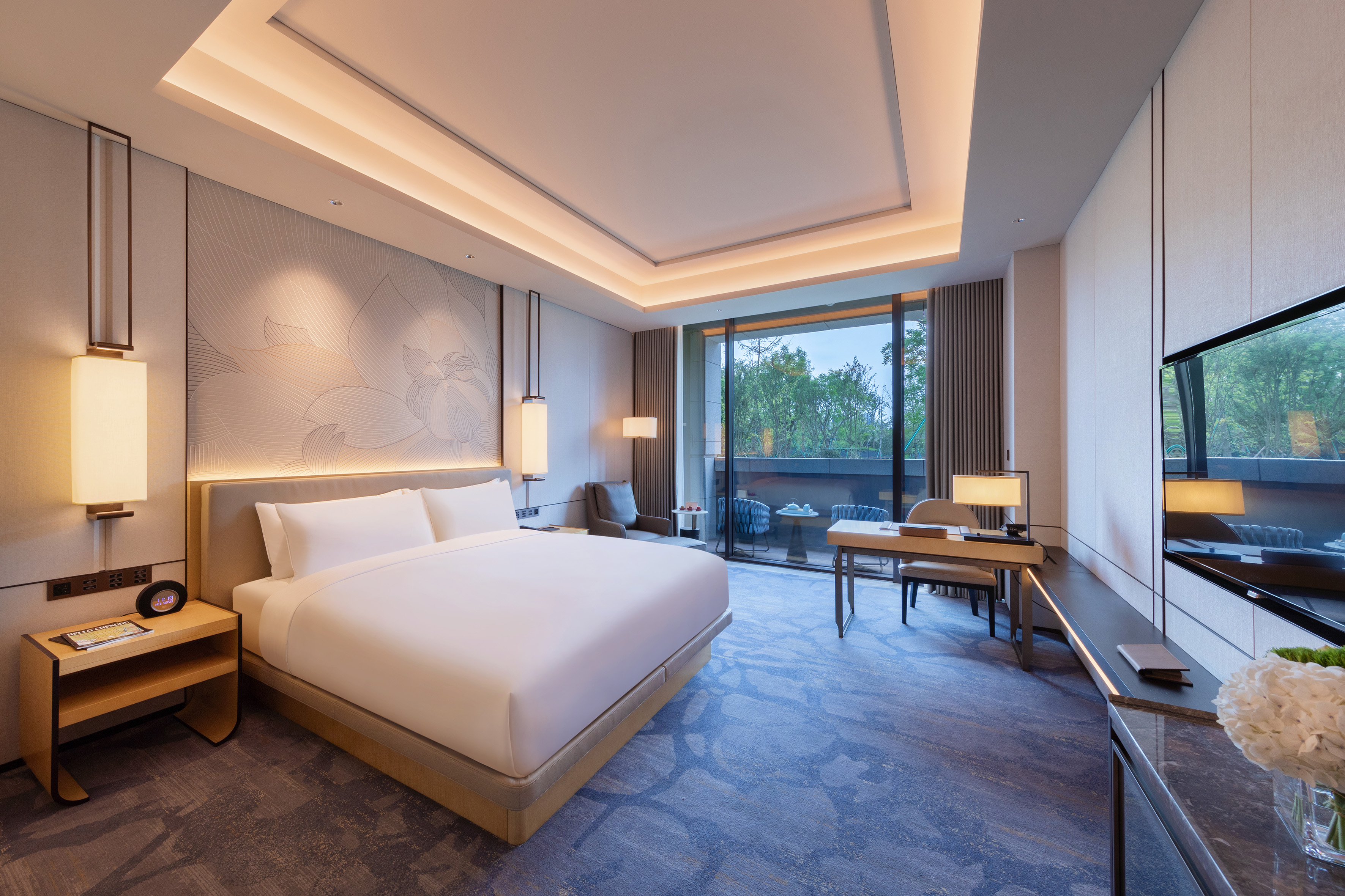 Modern hotel suite with king bed, floral wall, desk, and balcony.