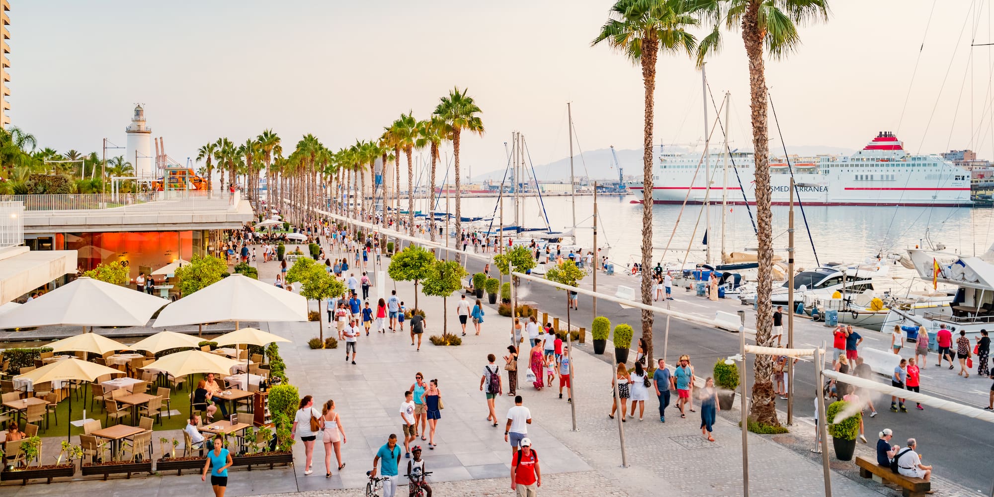 a group of people walking on a sidewalk with palm trees and boats