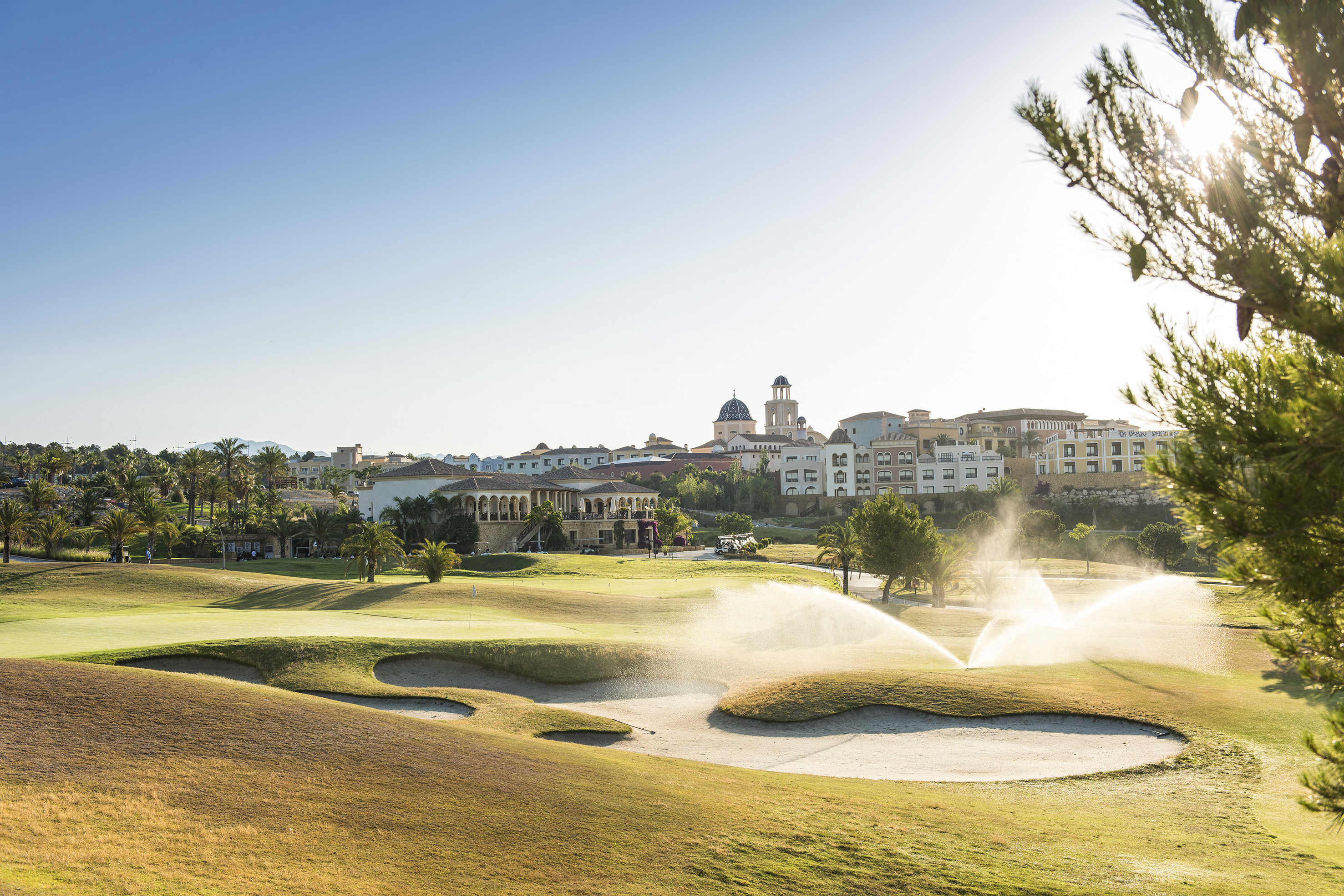 Sunny resort golf course with active sprinklers, bunkers, and distant buildings.