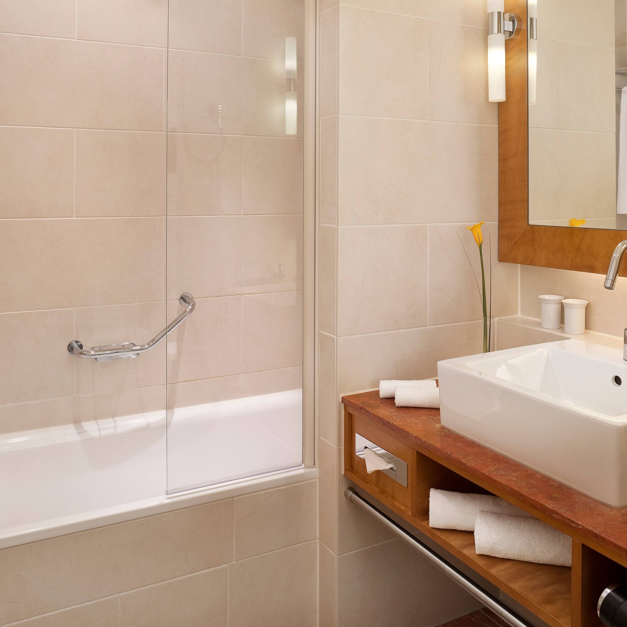 Modern hotel bathroom with bathtub, vanity sink, mirror, and beige tiles.