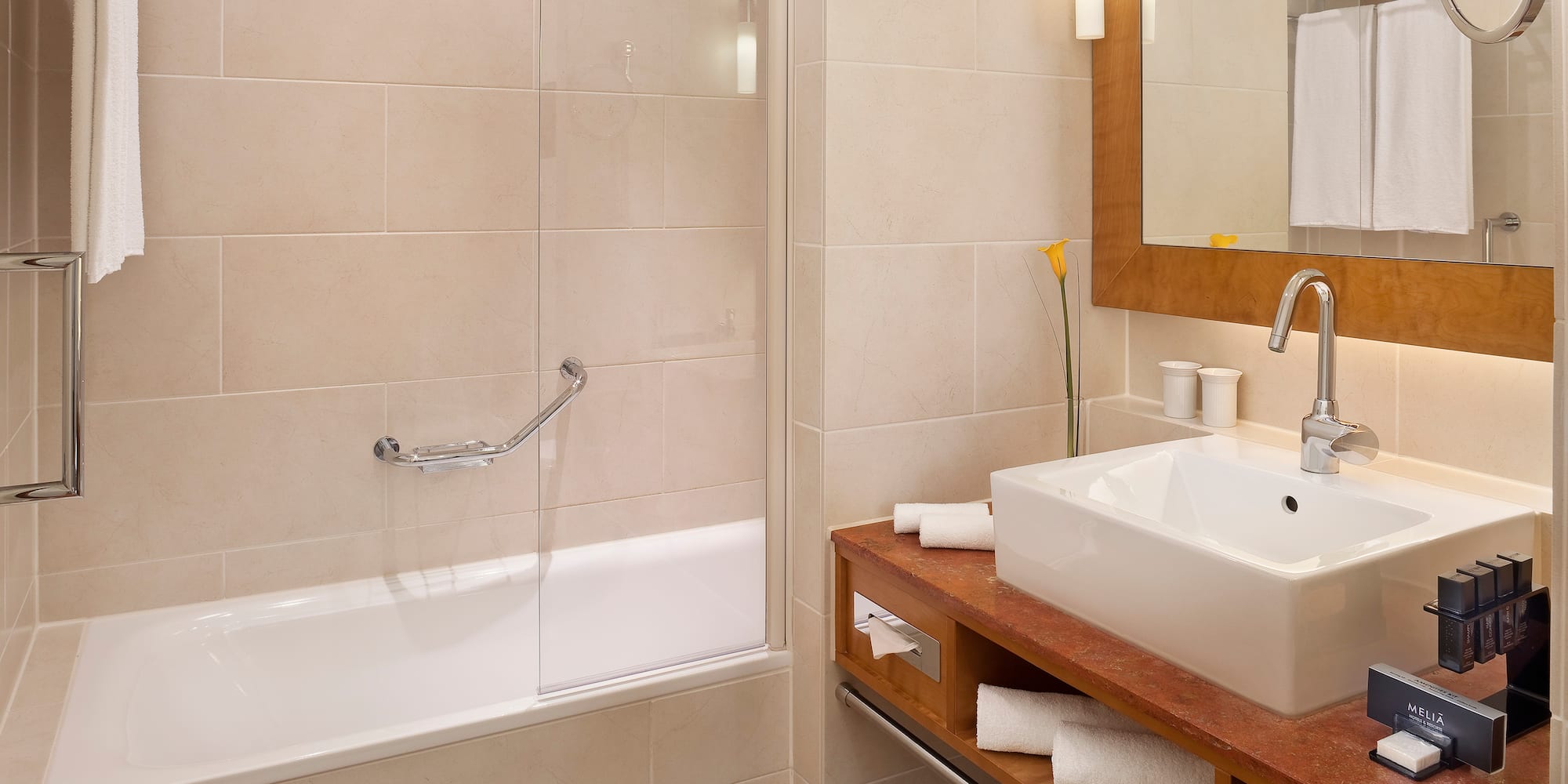 Modern hotel bathroom with bathtub, vanity sink, mirror, and beige tiles.