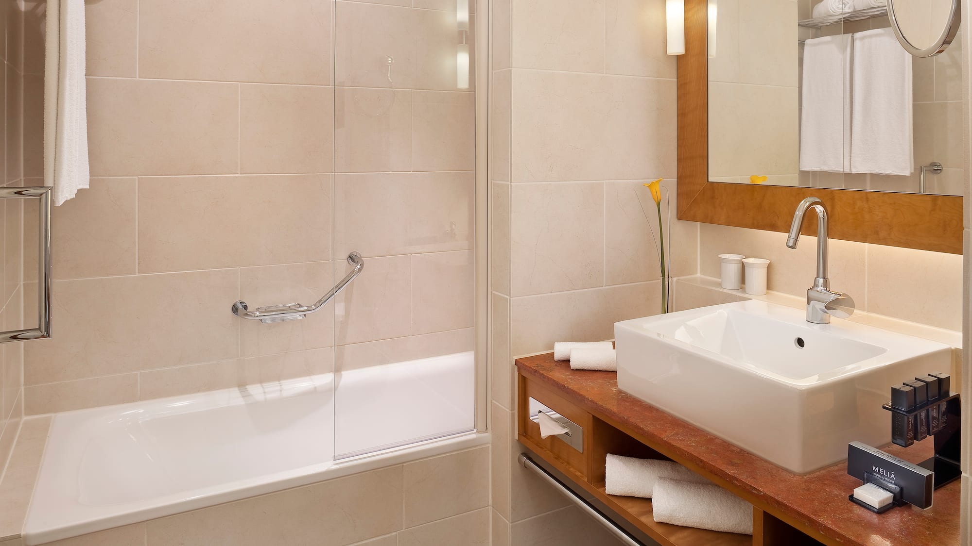 Modern hotel bathroom with bathtub, vanity sink, mirror, and beige tiles.