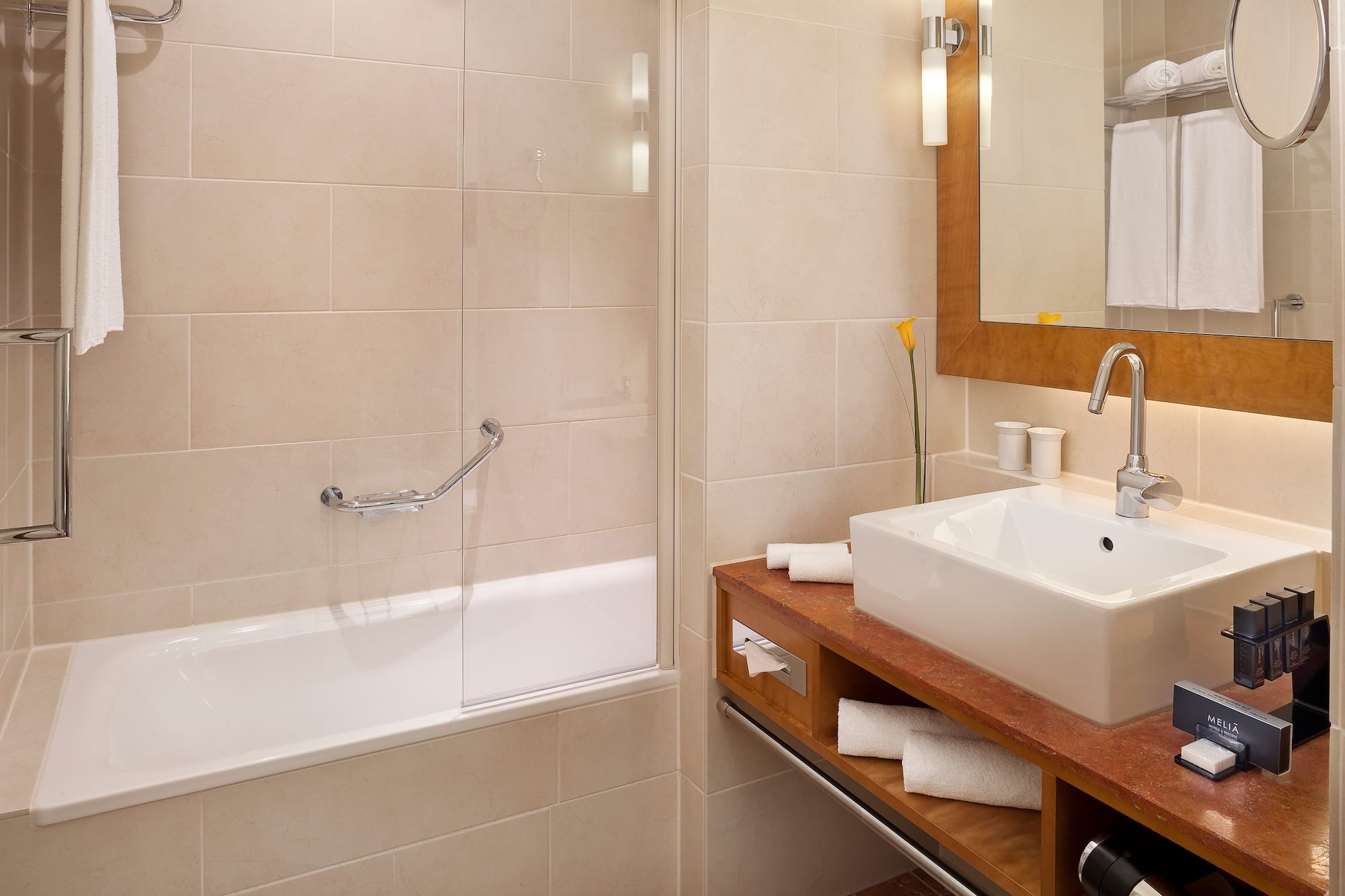 Modern hotel bathroom with bathtub, vanity sink, mirror, and beige tiles.