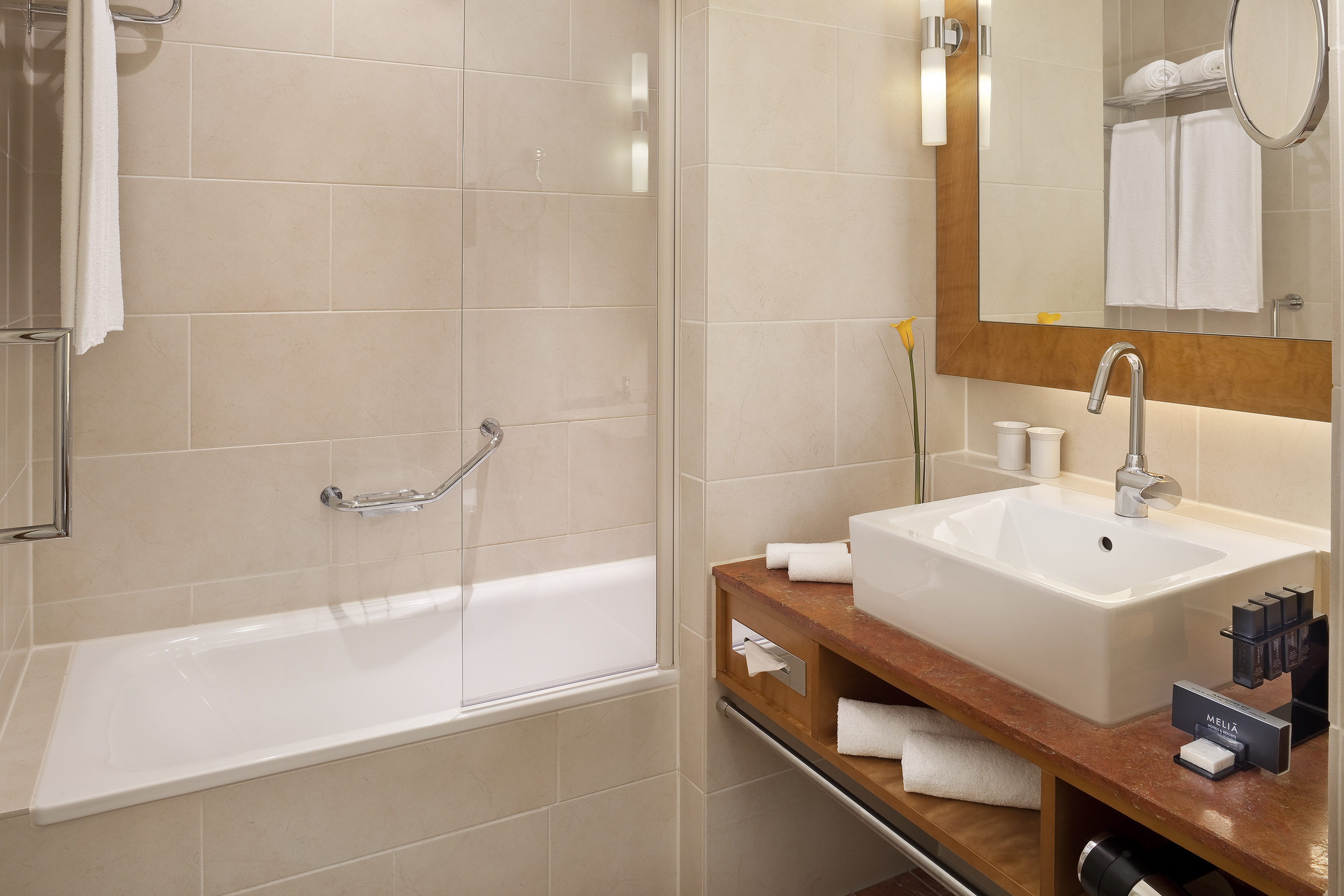 Modern hotel bathroom with bathtub, vanity sink, mirror, and beige tiles.