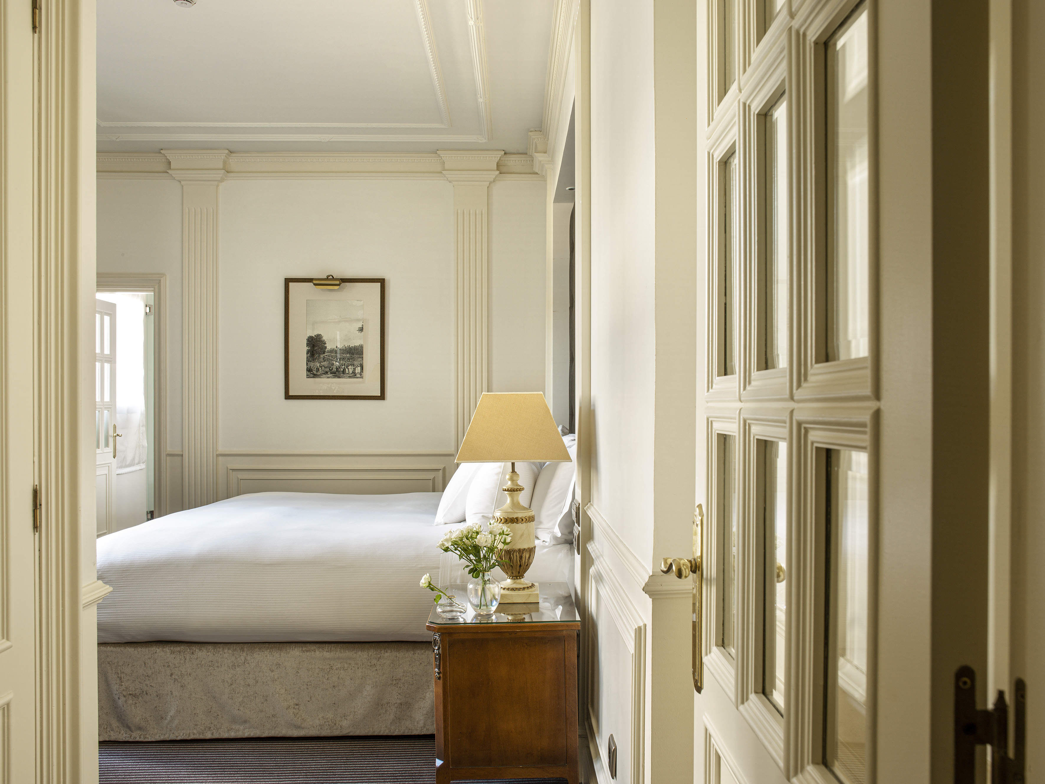 Elegant hotel bedroom with white bed, classic decor, nightstand, and ornate molding.