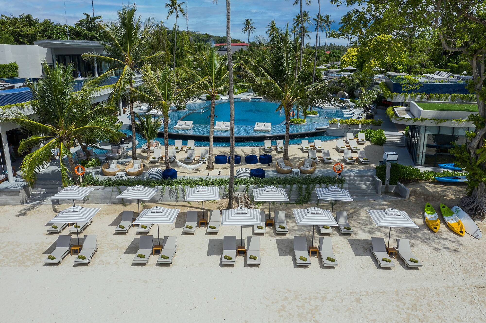 Luxury tropical hotel with large pool, palm trees, beach loungers on white sand.