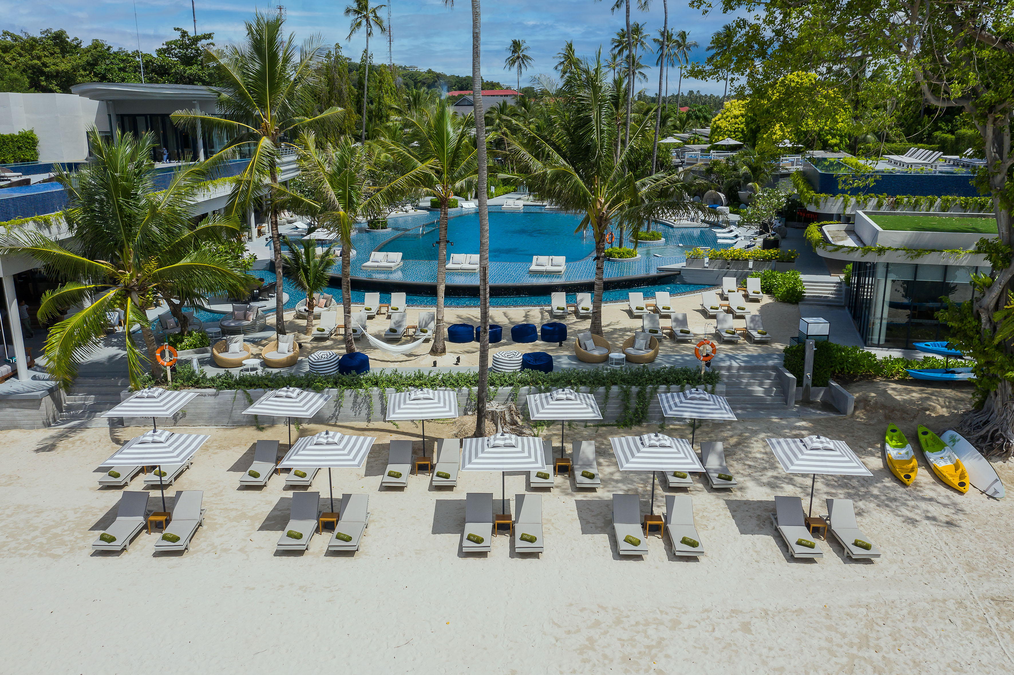 Luxury tropical hotel with large pool, palm trees, beach loungers on white sand.