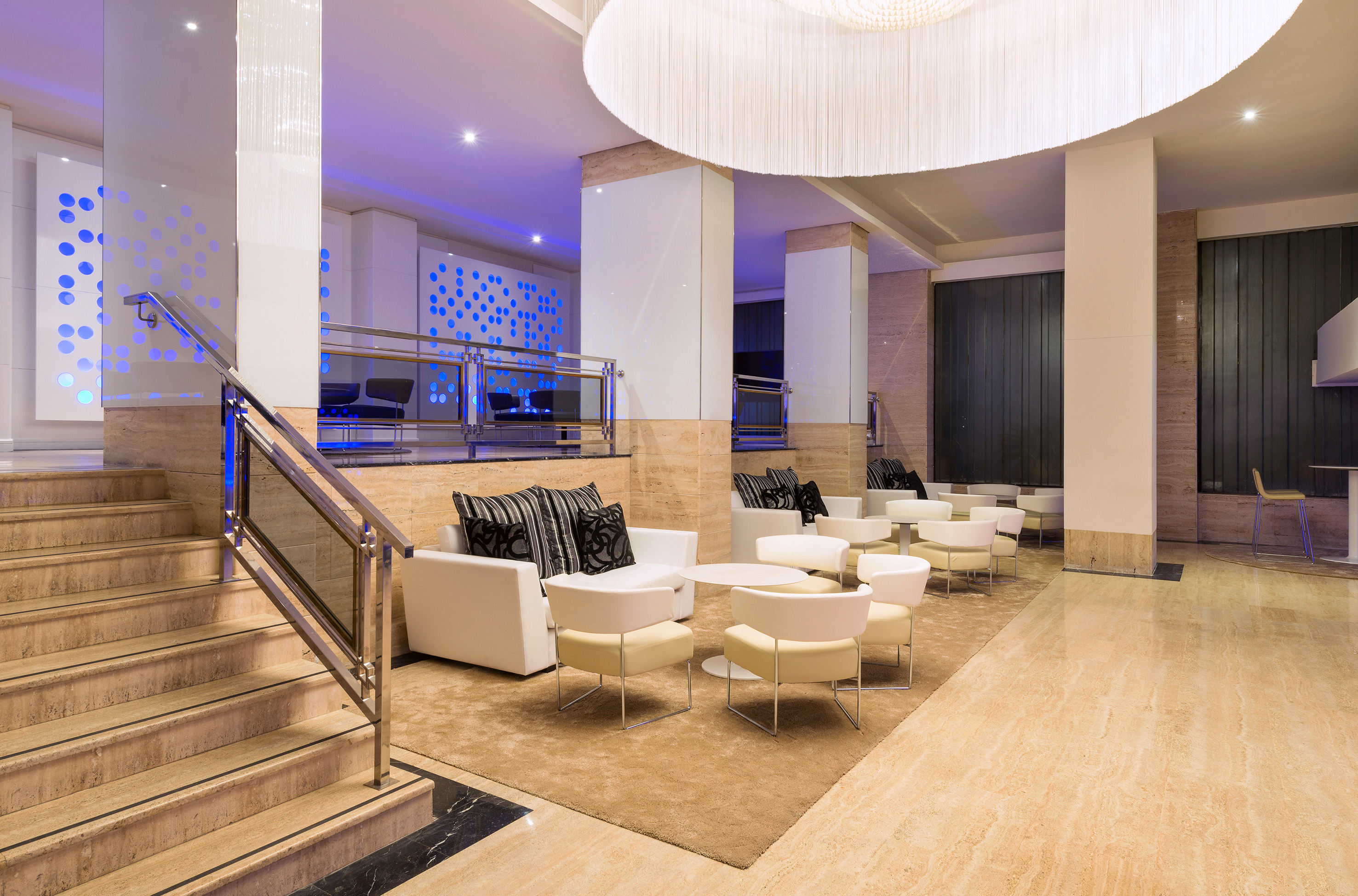 Modern hotel lobby lounge with white seating, marble stairs, and blue lighting.