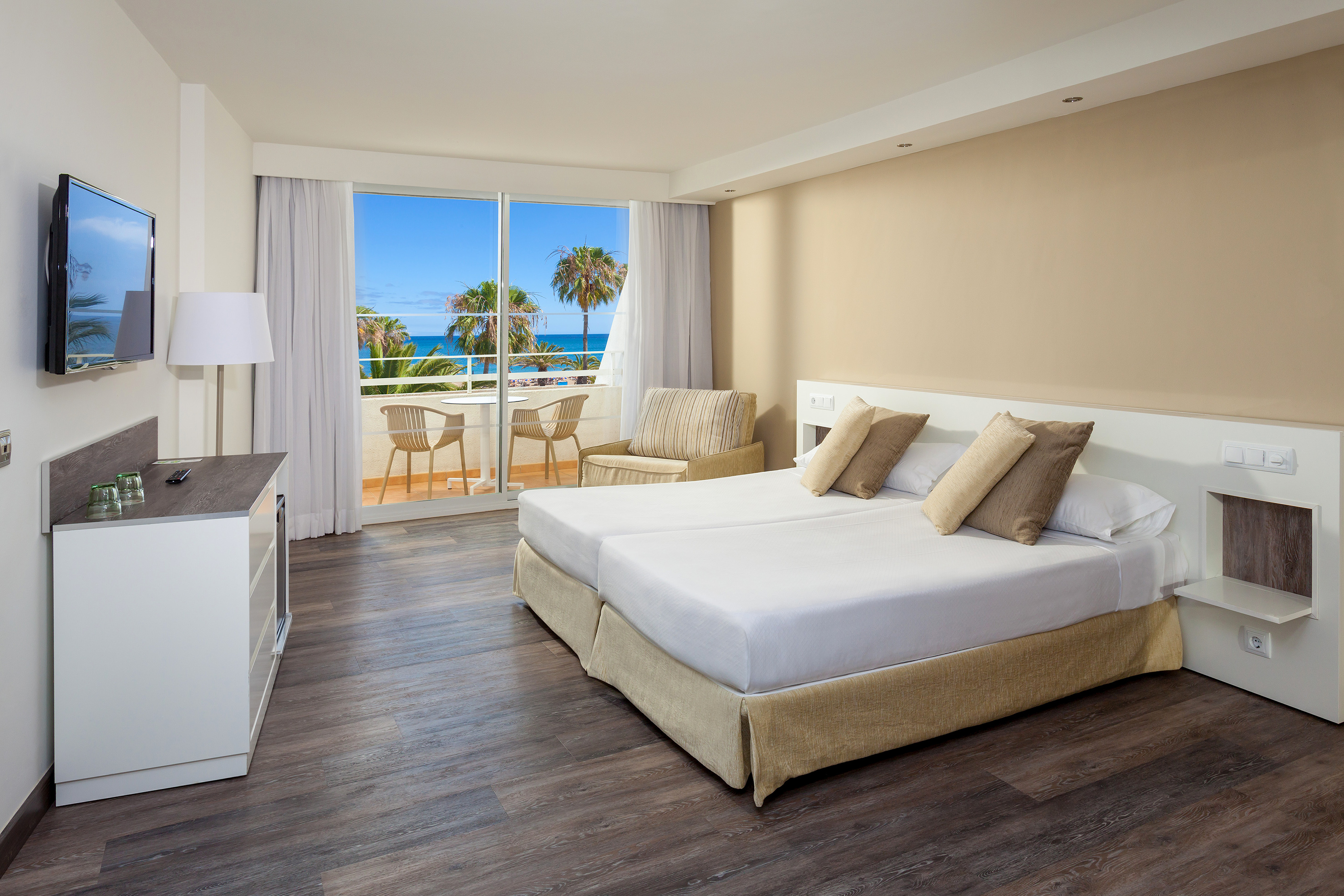Hotel room with ocean view balcony, twin beds, and tropical palm trees.