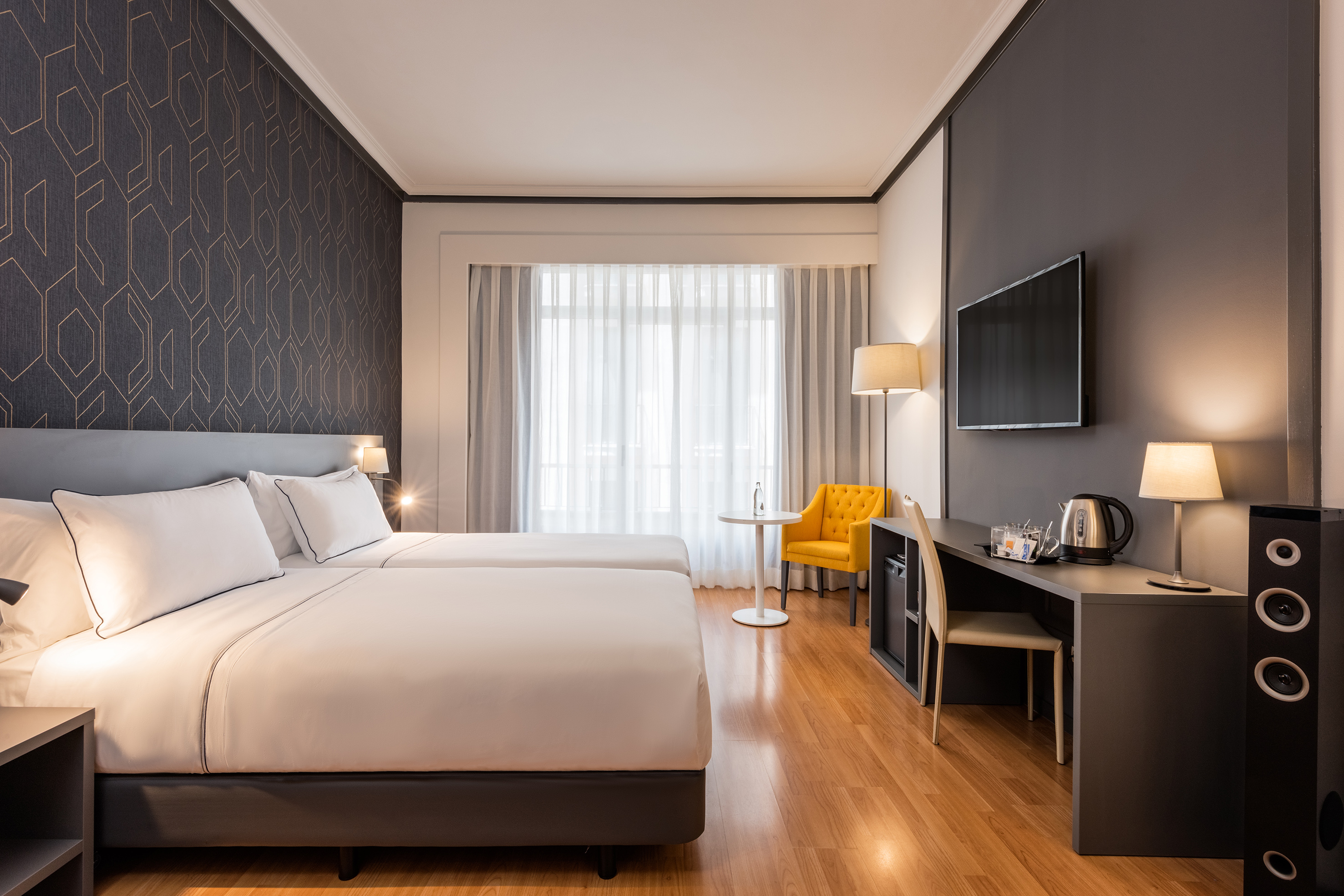 Modern hotel twin room with geometric wallpaper, desk, TV, and yellow armchair.