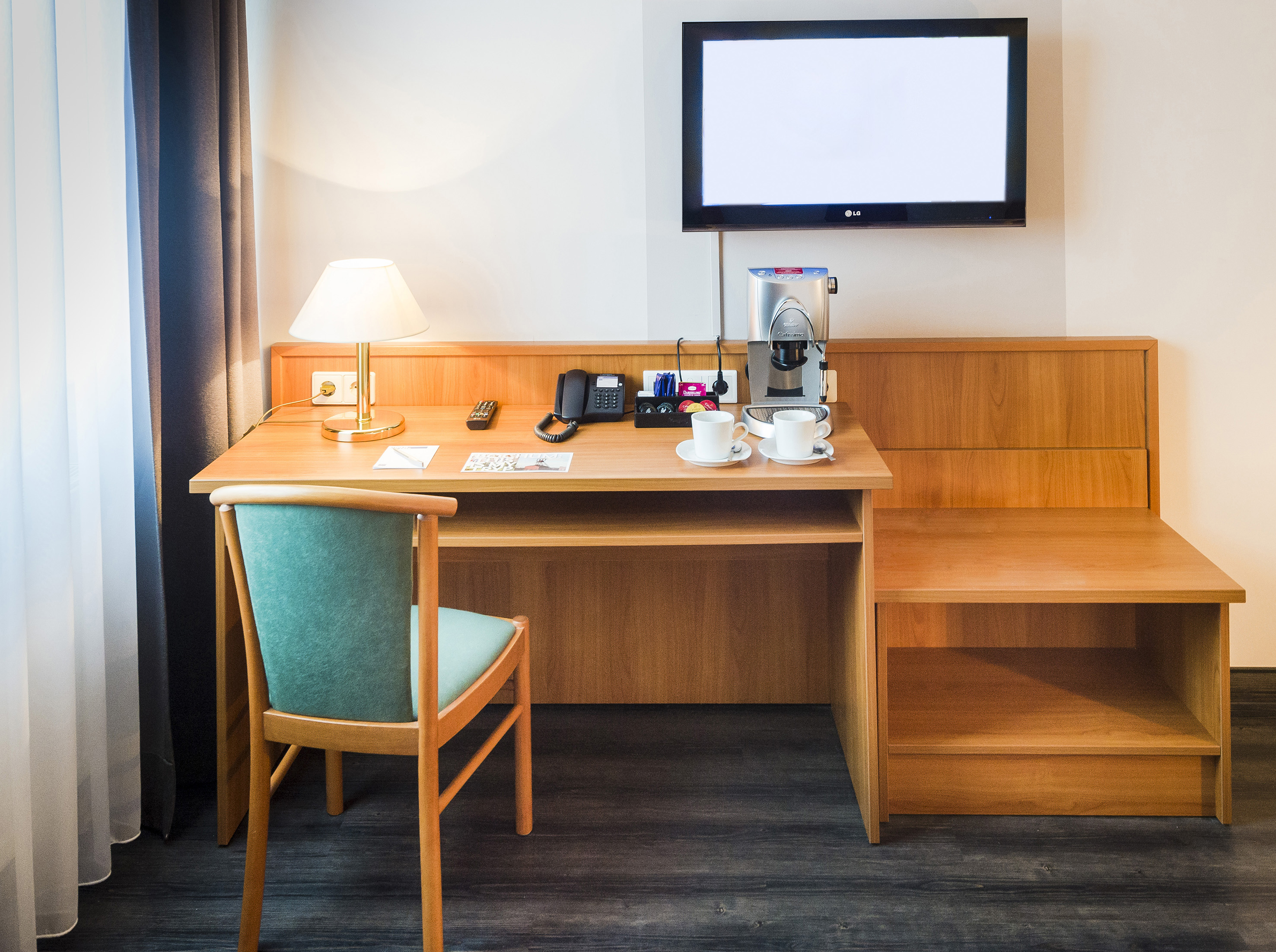 Hotel room workstation with wooden desk, green chair, wall TV, and coffee maker.