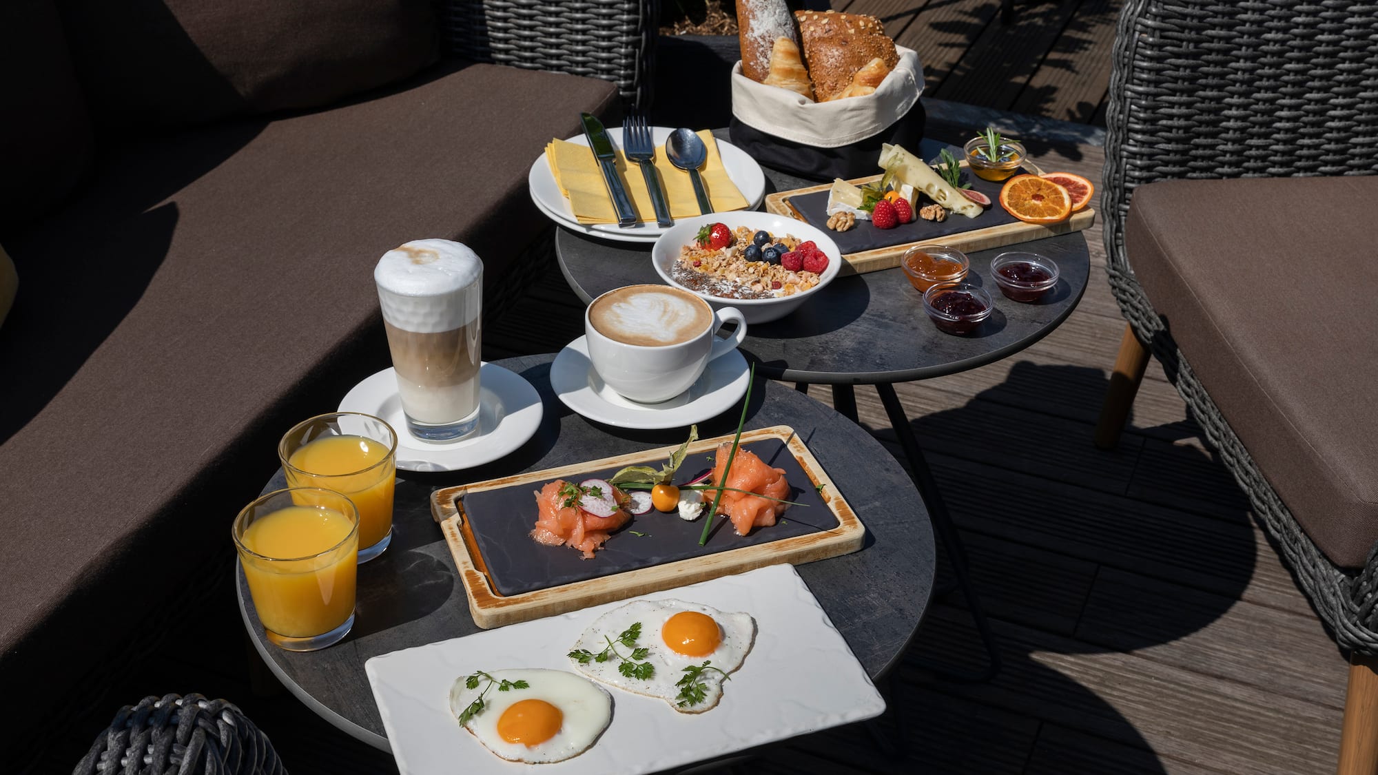 Sunny outdoor breakfast: coffee, eggs, smoked salmon, granola, bread, cheese, juice.
