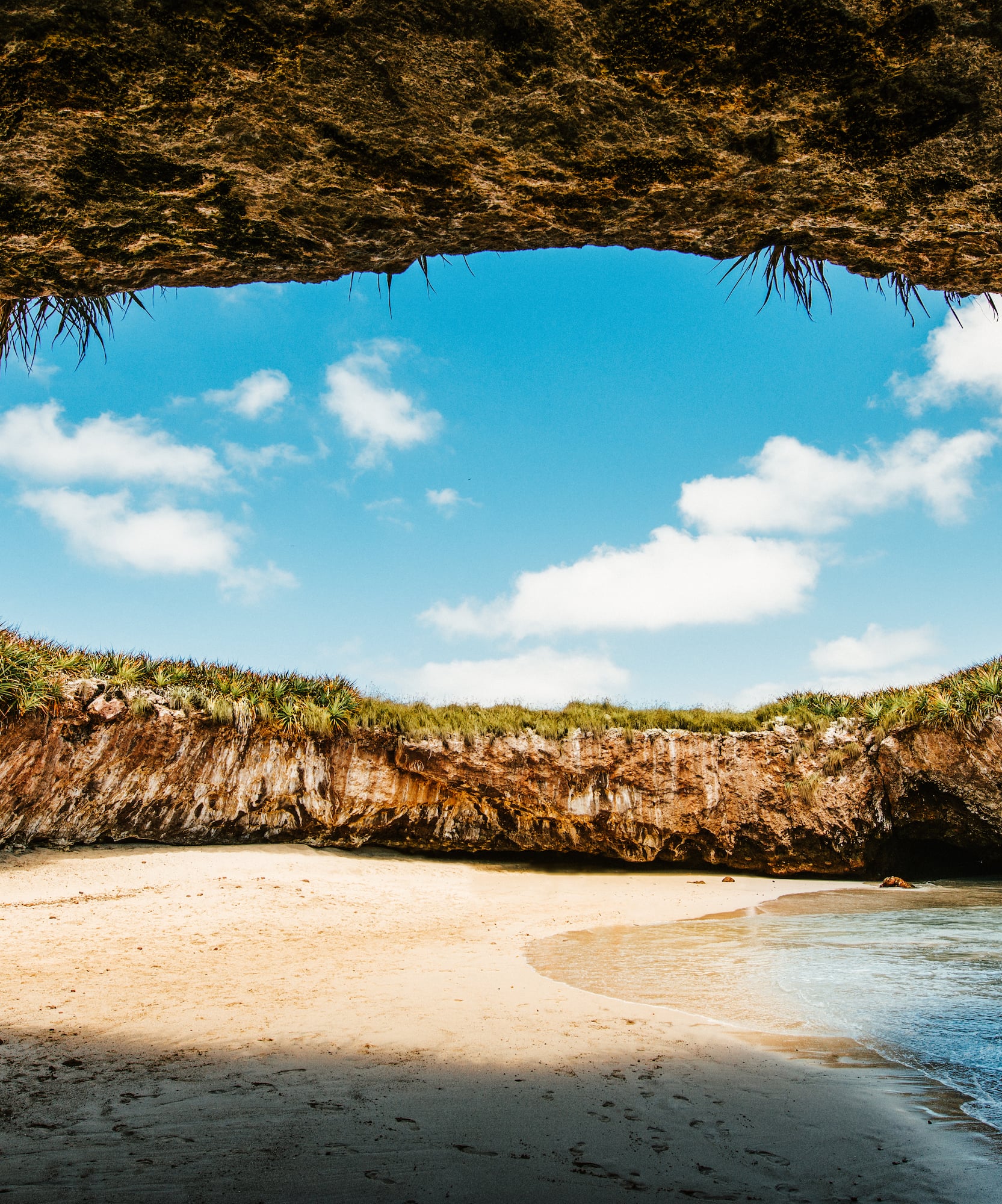Cueva rocosa, playa de arena con agua turquesa y cielo azul.
