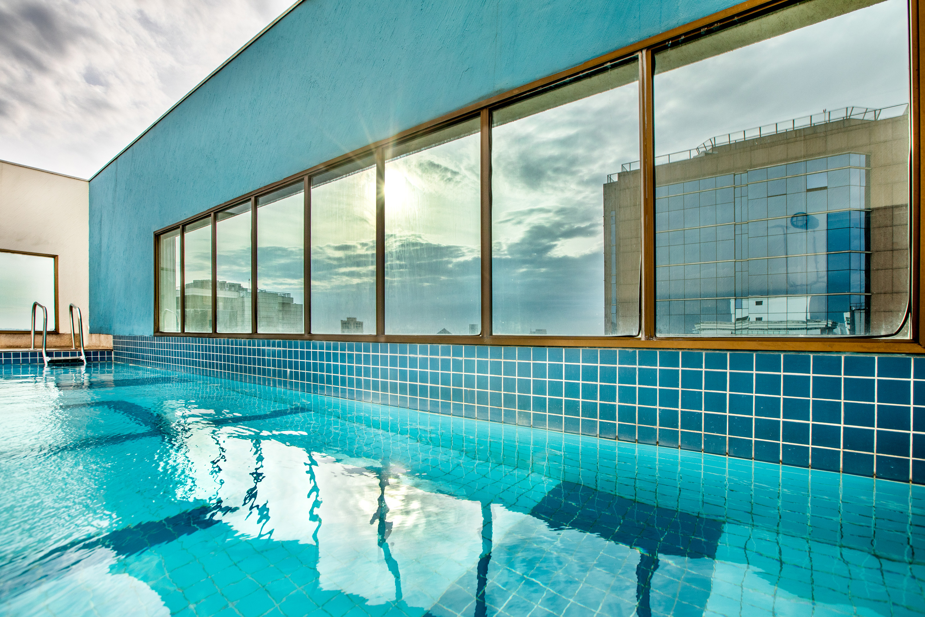 Modern rooftop swimming pool with blue tiles, windows reflecting city skyline.