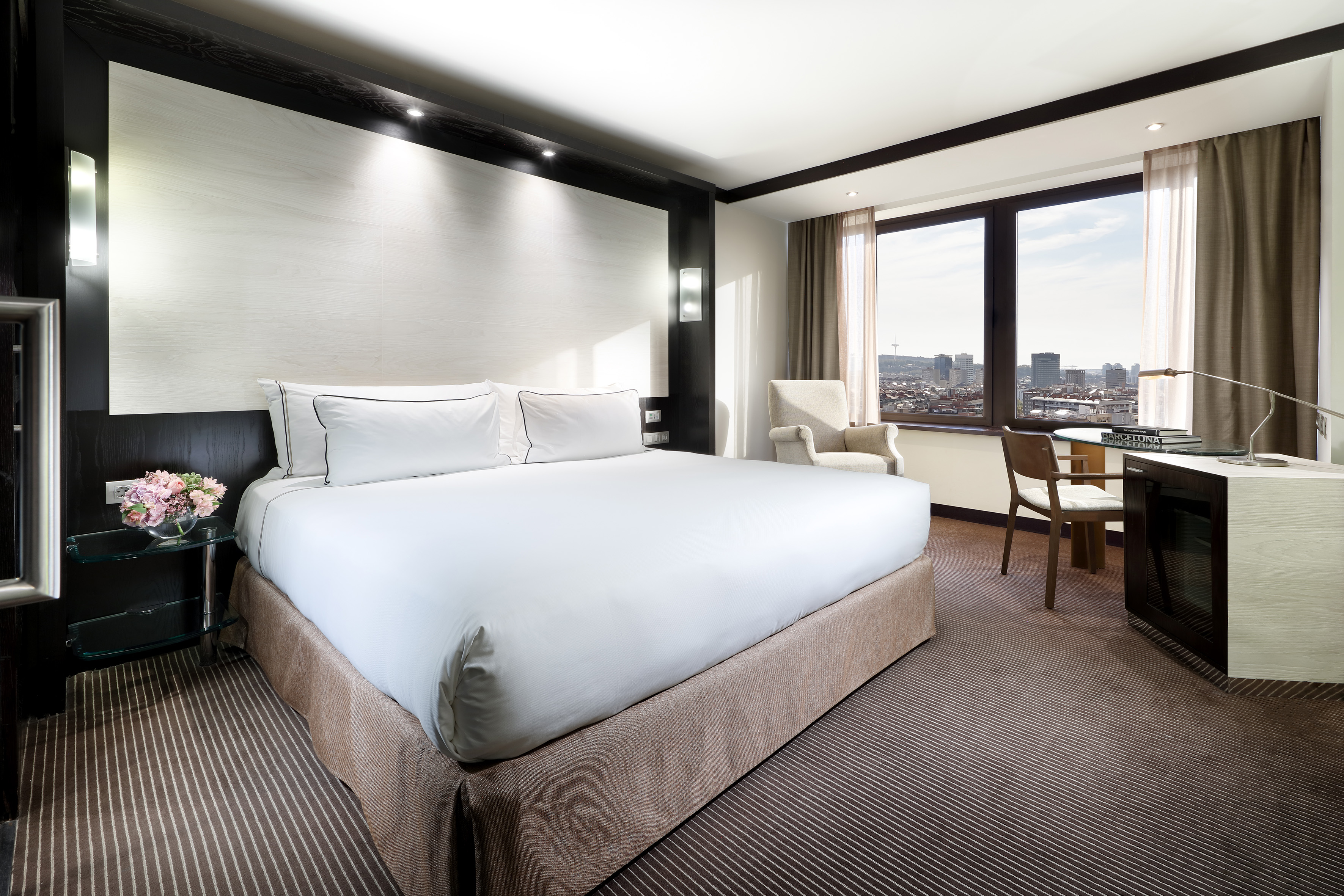Modern hotel room with white bed, cityscape view, and work desk.