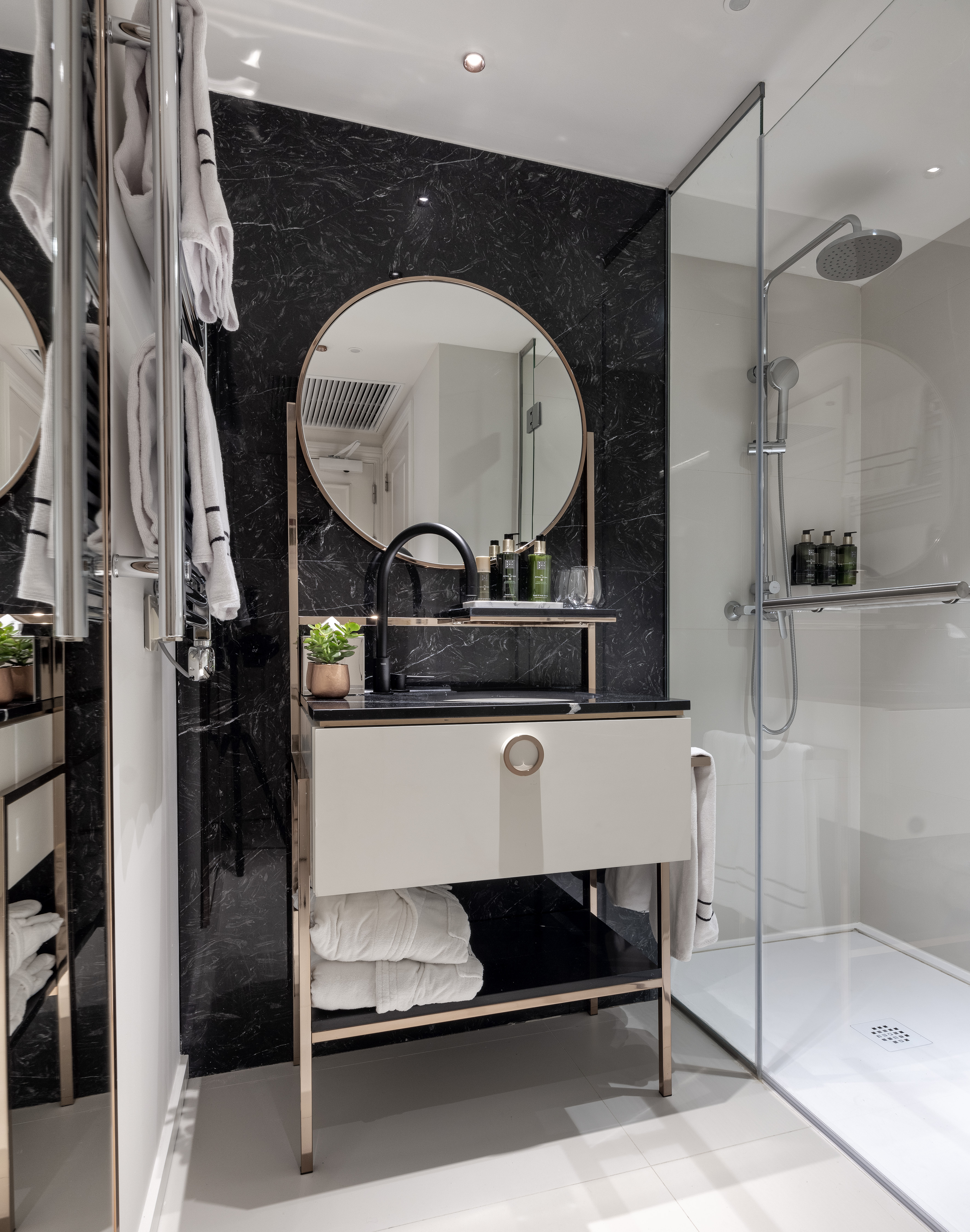 Modern bathroom with black marble, round mirror, gold vanity, and glass shower.