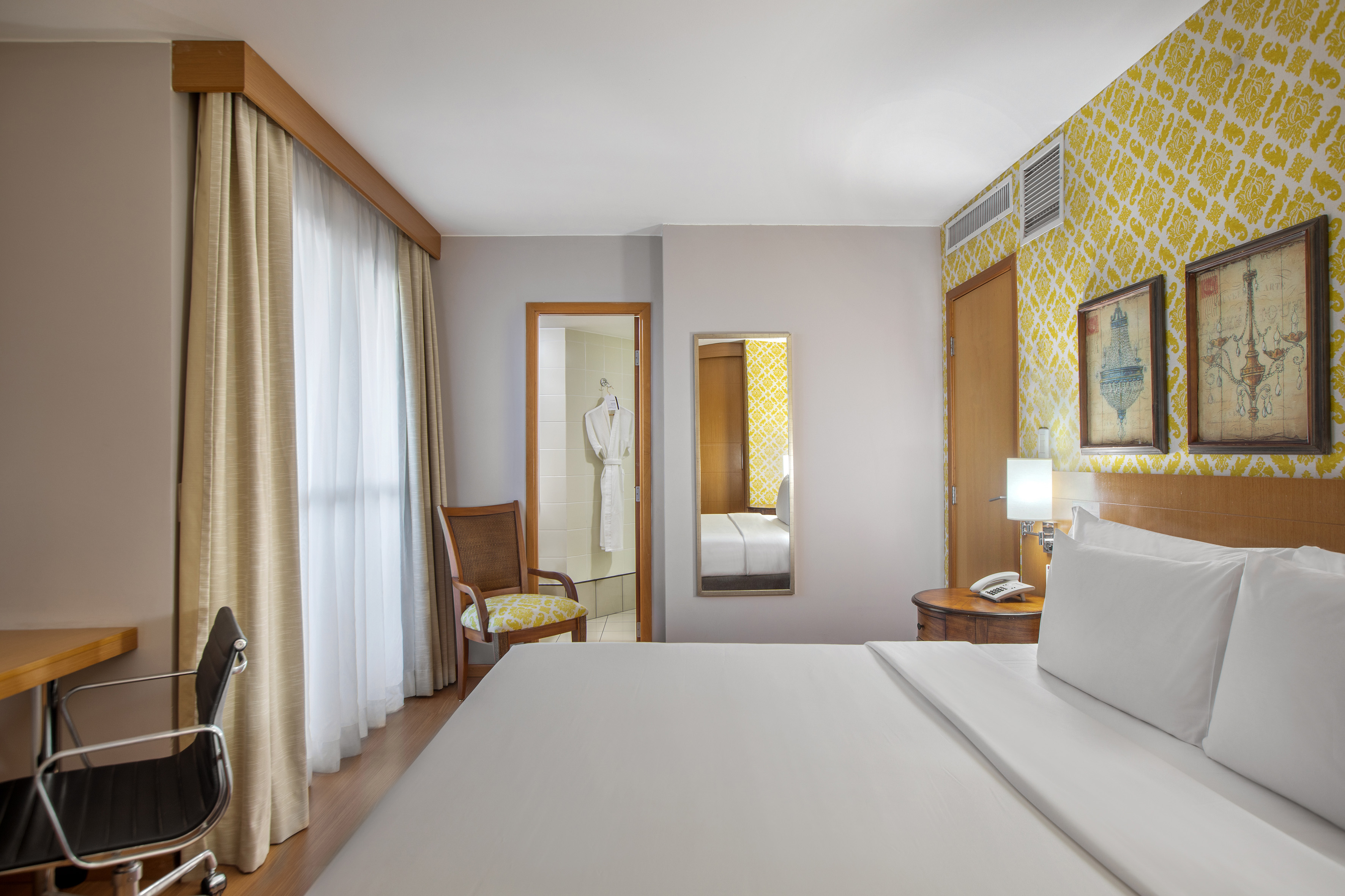 Modern hotel room with king bed, bright yellow accent wall, desk, and bathrobes.