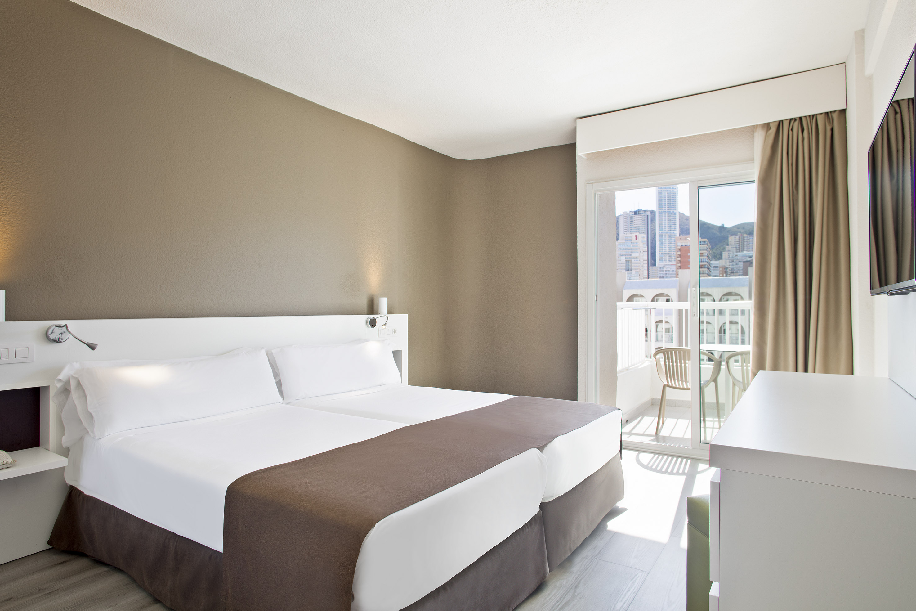 Hotel room with twin beds, brown wall, balcony, and city view.
