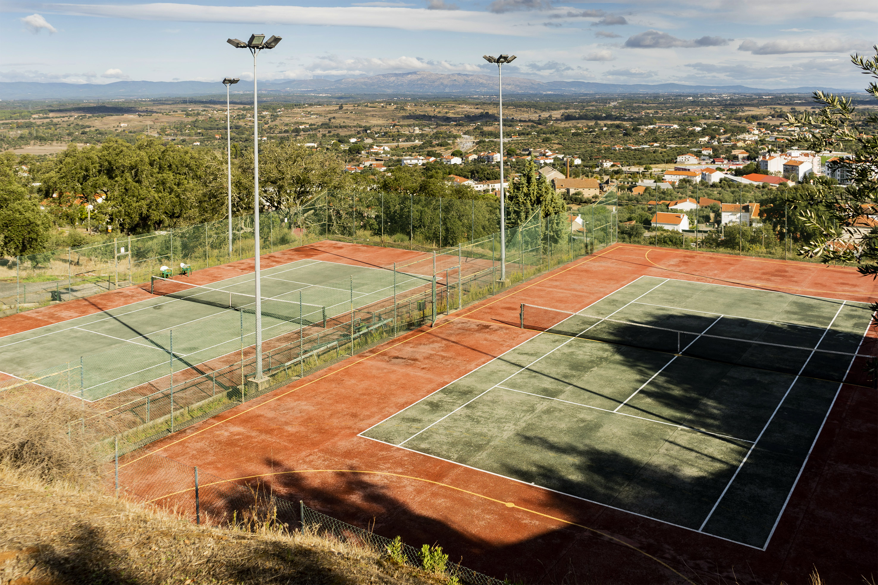 Two outdoor tennis courts with floodlights, village, and distant mountains in background.