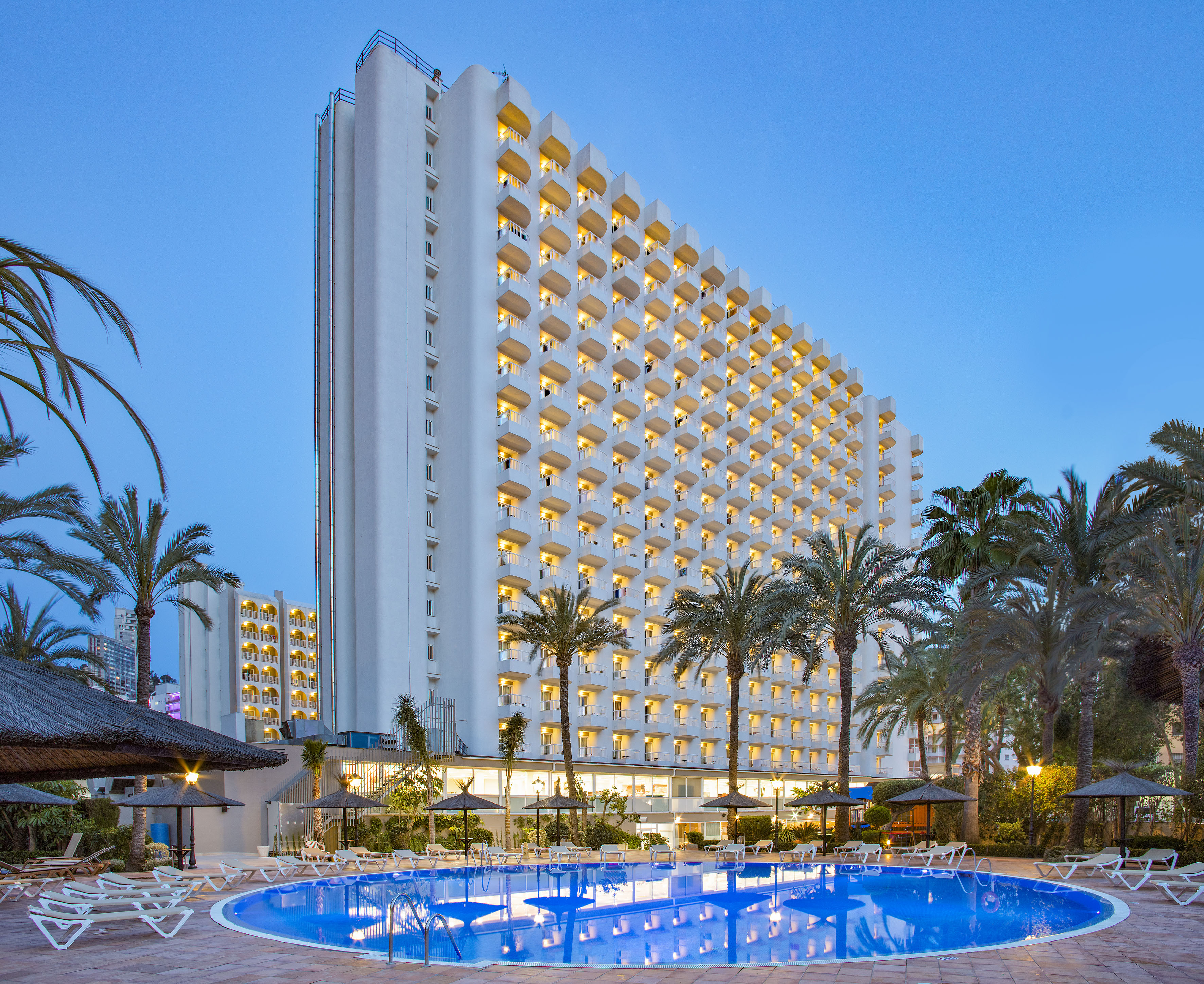 Illuminated hotel resort with swimming pool and palm trees at dusk.