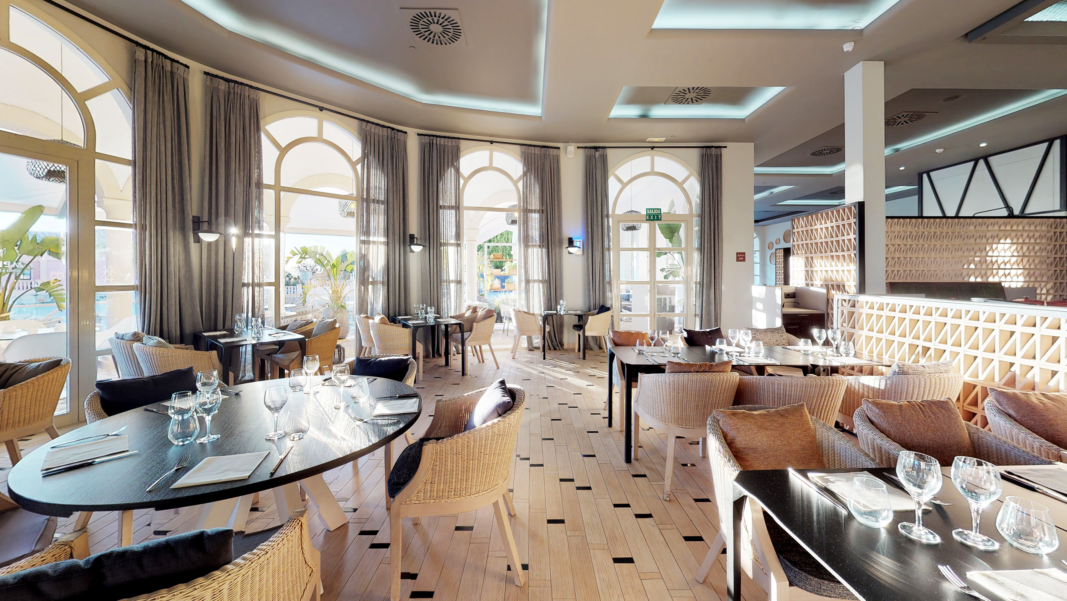 Sunny resort restaurant with arched windows, wicker chairs, and set dining tables.