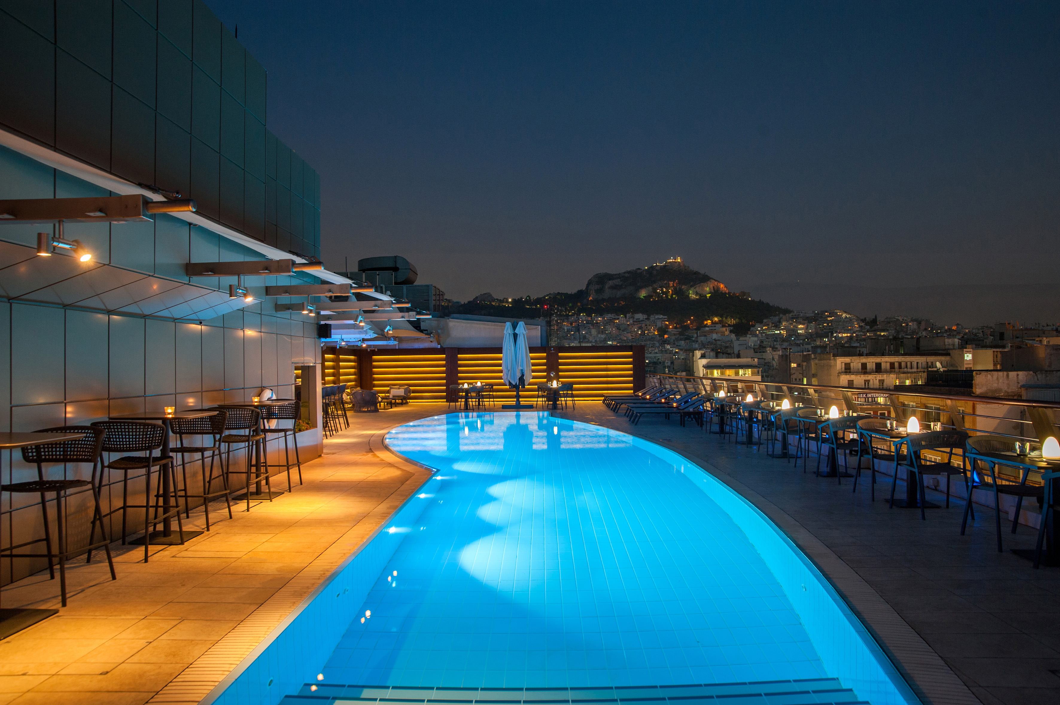 Rooftop pool bar with illuminated city skyline and Acropolis at night in Athens.