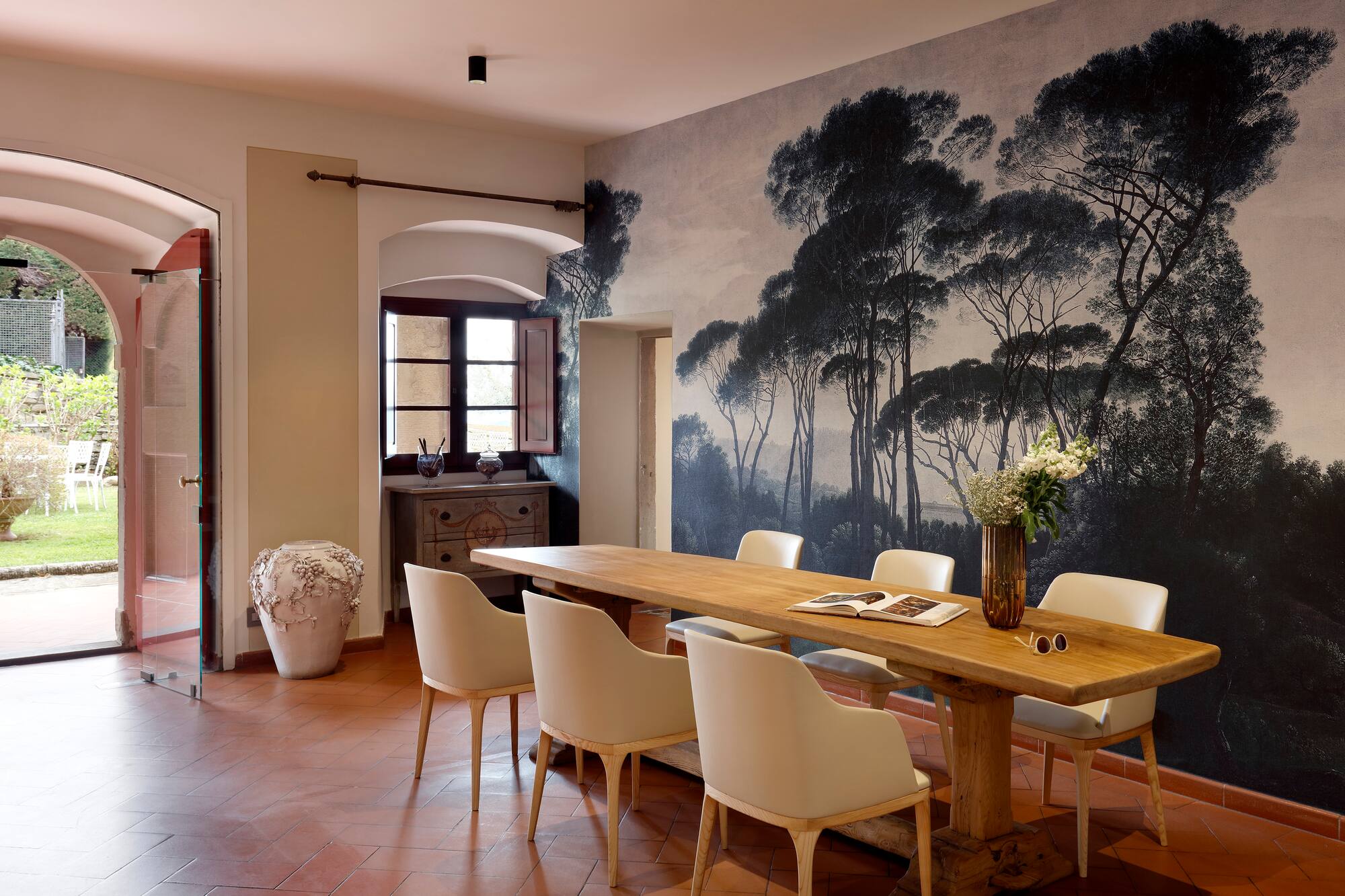 Dining room with long wooden table, beige chairs, forest mural, and terracotta floor.