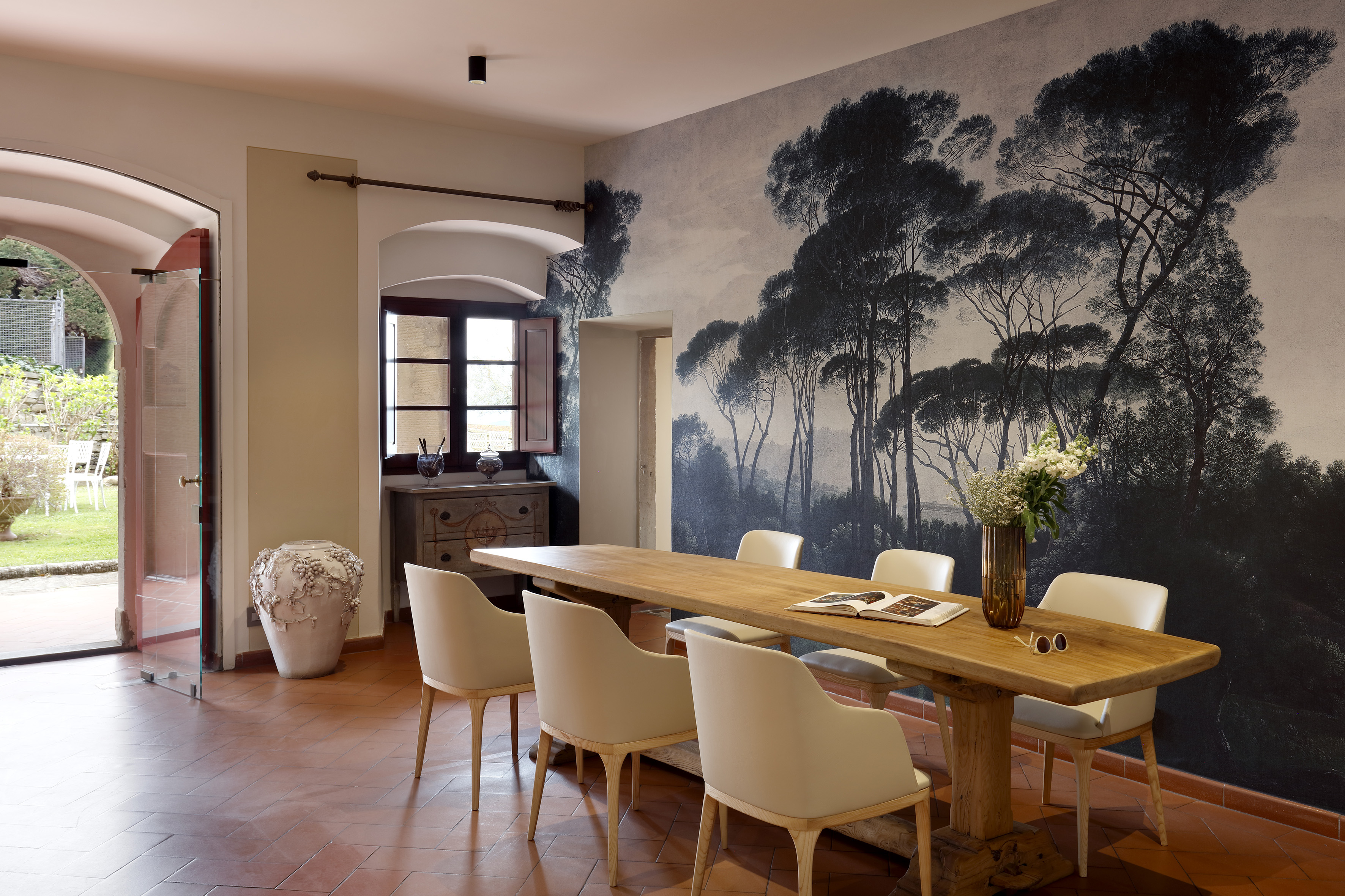 Dining room with long wooden table, beige chairs, forest mural, and terracotta floor.